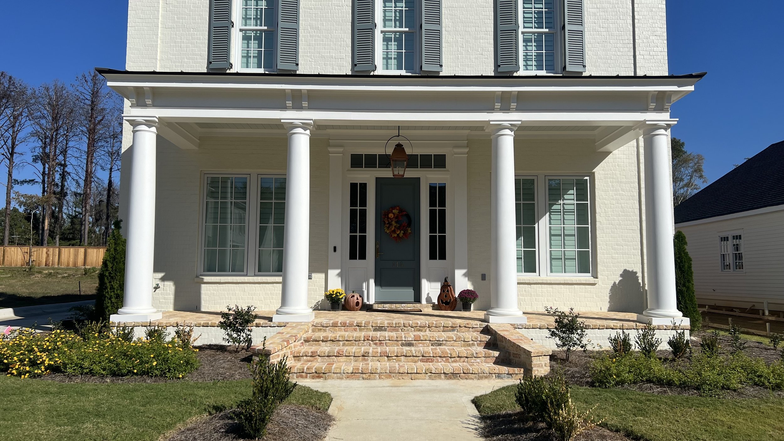 Front view of a white two-story house with a porch supported by four white columns, brick stairs, and a teal front door decorated with a fall-themed wreath. Decor on the porch includes pumpkins, potted flowers, and a lantern, with a well-maintained g