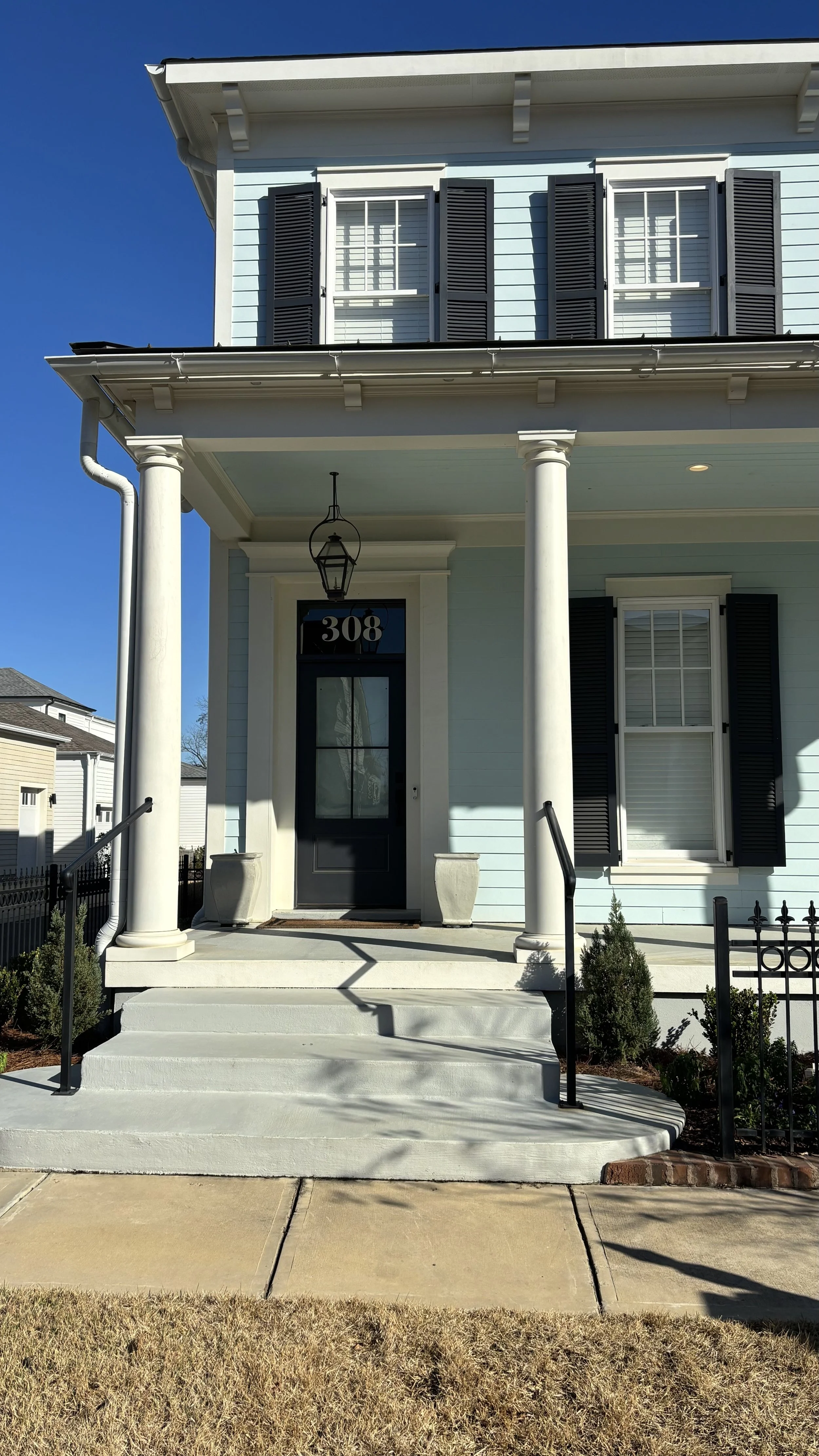 Front view of a house with light blue siding, white trim, black shutters, and black door with house number 308. The house has a porch with white columns, stairs, and potted plants, and is under a clear blue sky.
