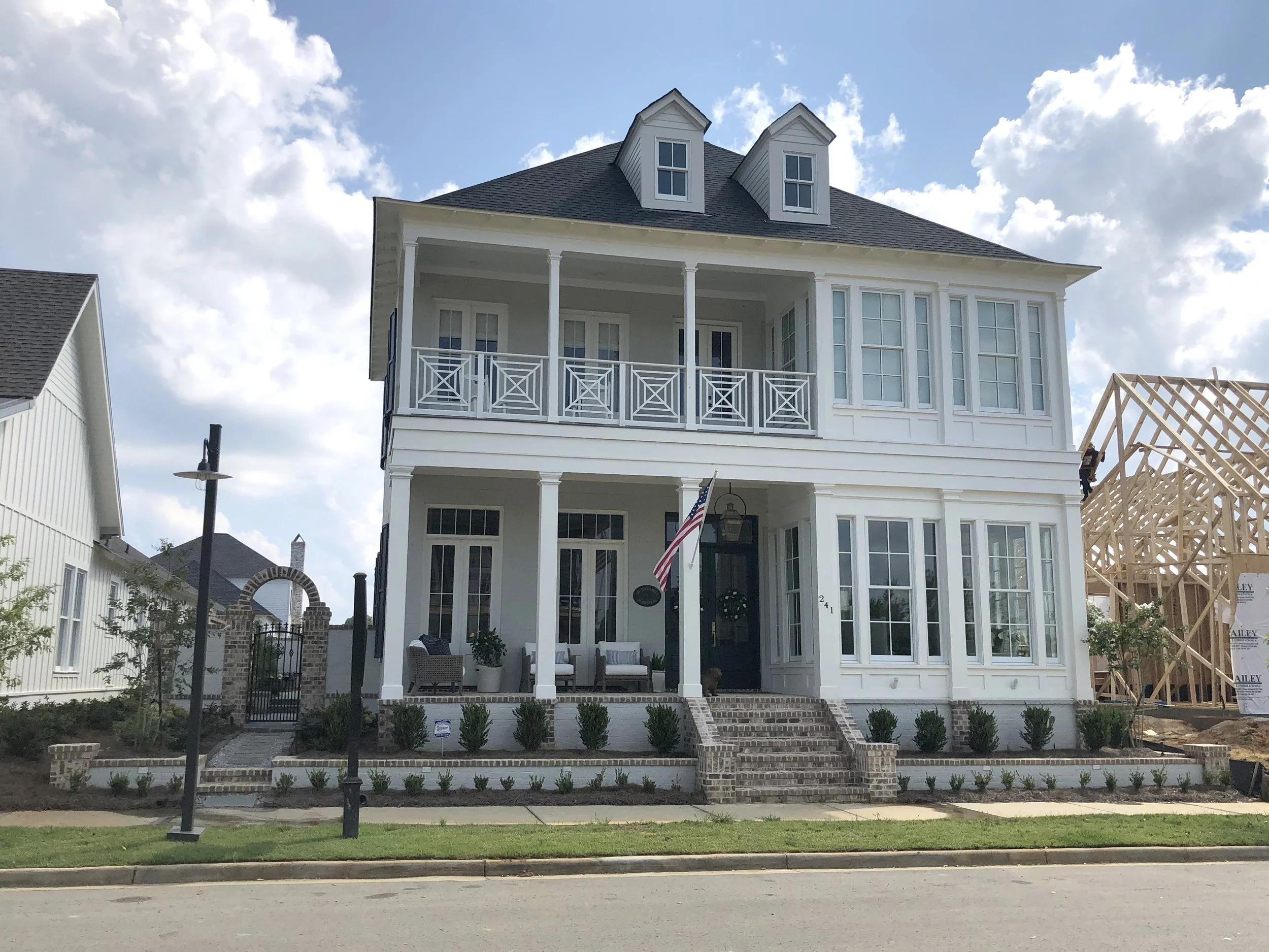 A newly constructed white three-story house with large windows, front porch, and a decorative balcony. There is a small garden with bushes and a lawn in front, and a construction site with wooden framing is visible on the right side.