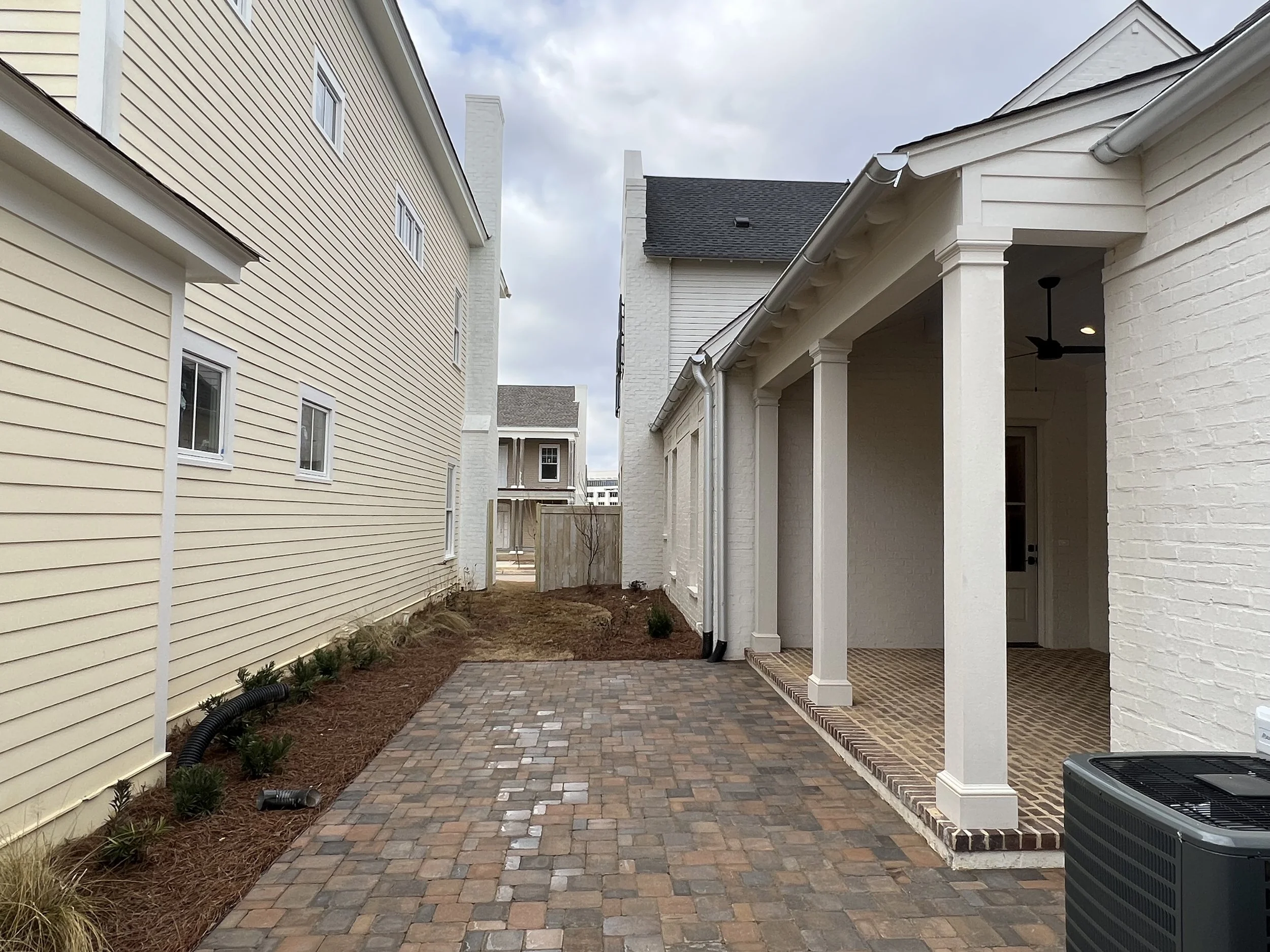 View of a narrow backyard area between two houses with brick pavers, a small porch with white columns, and a fence in the distance under an overcast sky.
