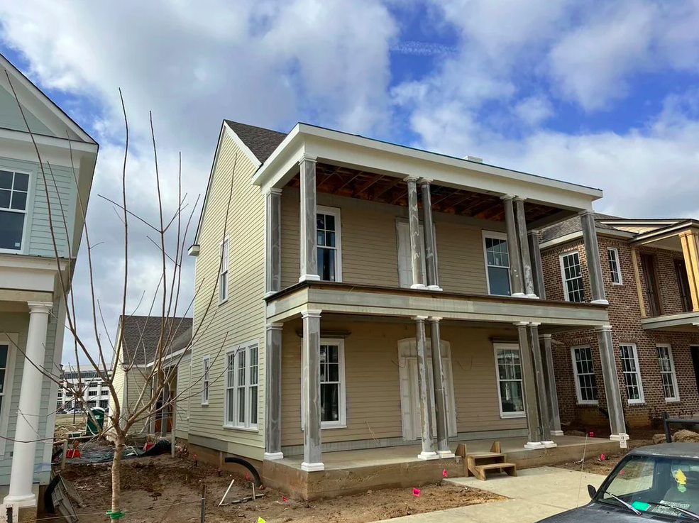 A two-story house under construction with a beige exterior, white trims, and columns supporting the porch and balcony. The front yard is unfinished with dirt and construction materials. Other houses are visible nearby, and the sky is partly cloudy.