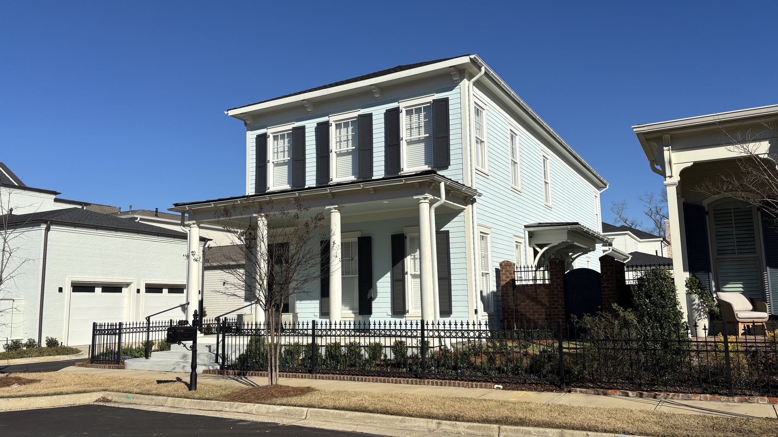 A light blue, two-story house with black shutters, white trim, and a front porch supported by white columns. The house is surrounded by a black metal fence, with small trees and shrubs in the front yard. The sky is clear and blue.