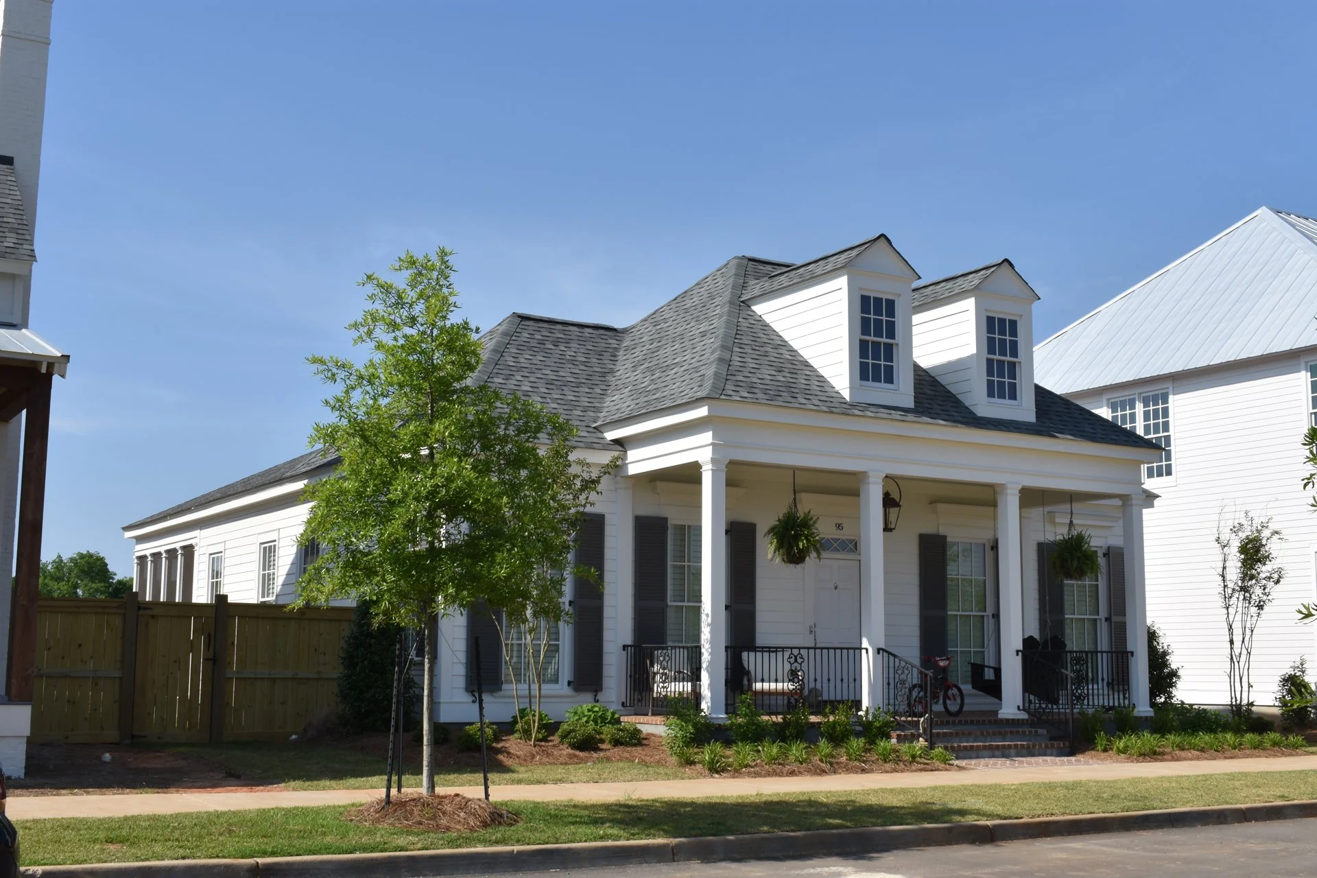 White two-story house with a front porch, black shutters, two dormer windows on the roof, and hanging plants, surrounded by a lawn and young trees.
