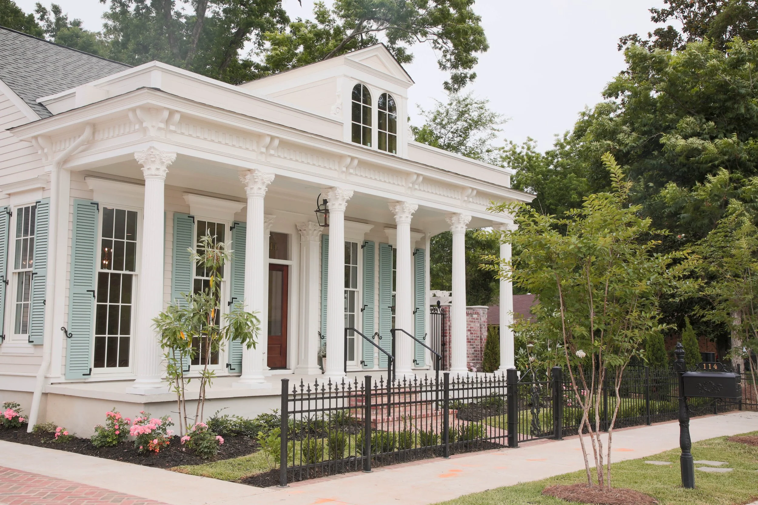 White house with light blue shutters, columns at the porch, steps leading up to the door, surrounded by a small garden with pink flowers and trees, black fence and mailbox in front.
