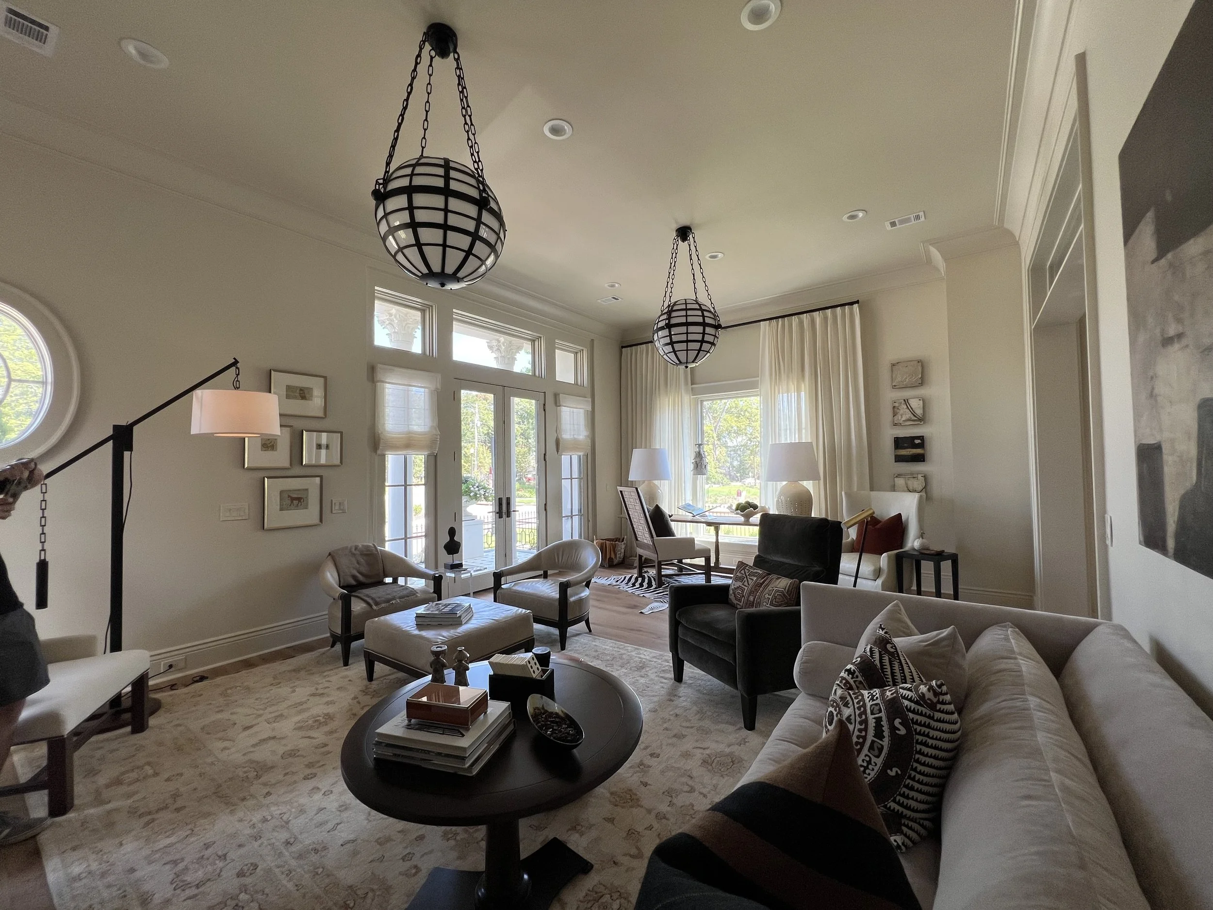 Luxurious living room featuring two large, spherical pendant lights hanging from the ceiling, cream-colored sofas with decorative pillows, a dark wood coffee table with books and decorative items, multiple armchairs, and large windows with white curt