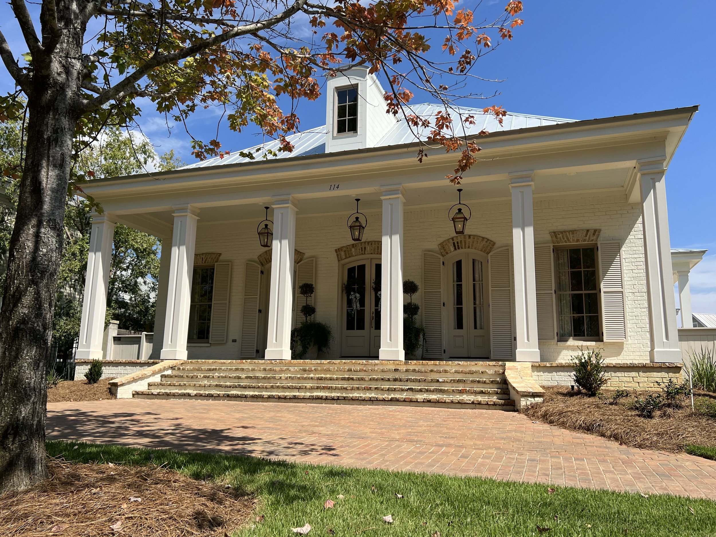 Front view of a white, two-story house with a large front porch supported by columns, brick stairs, and a brick walkway. The house has a metal roof and several windows with shutters. There are trees and landscaped bushes in the yard, and the sky is c