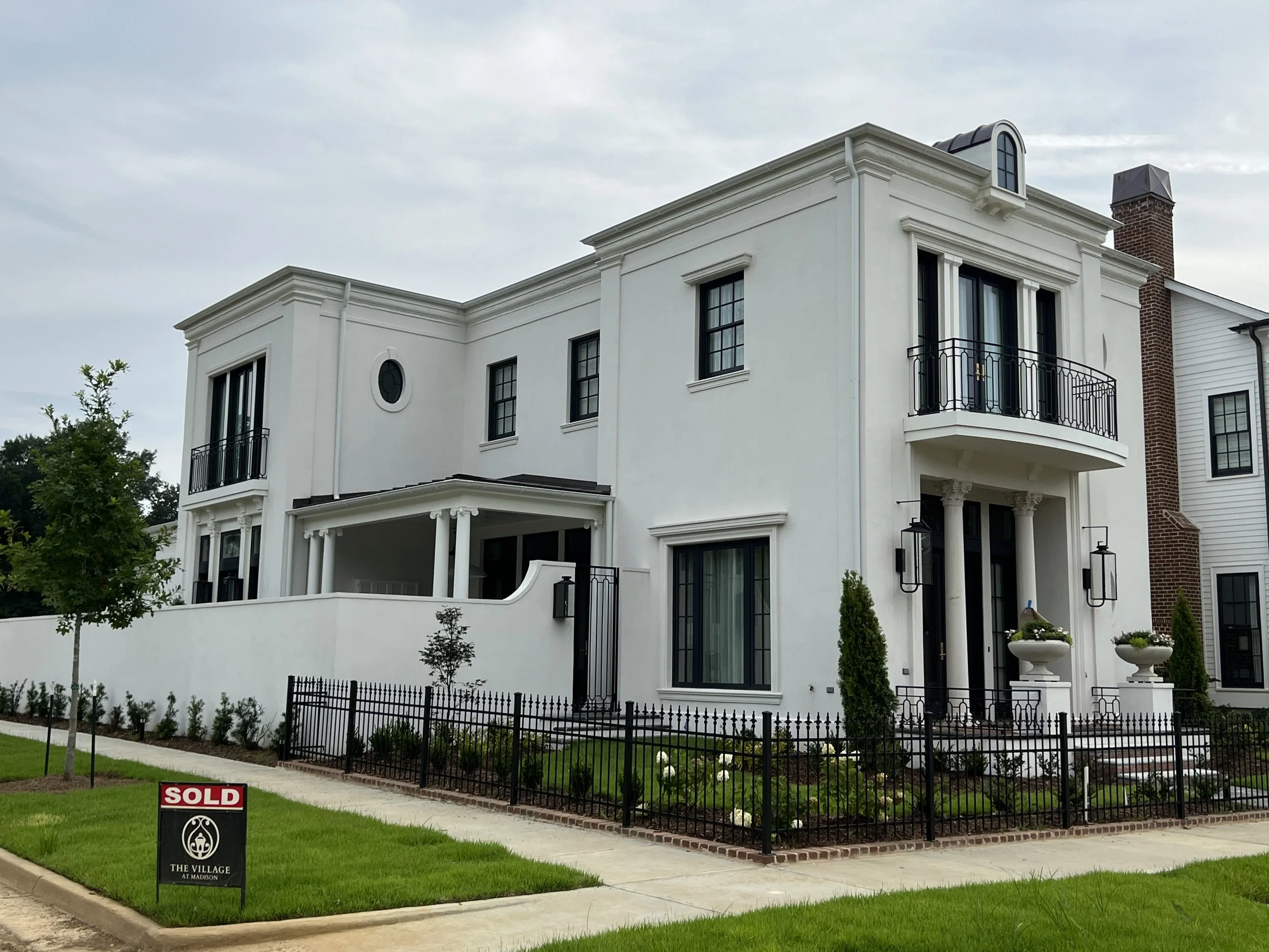 White multi-story house with black window frames and decorative columns, front yard with black wrought iron fence, and a 'Sold' sign in the lawn.