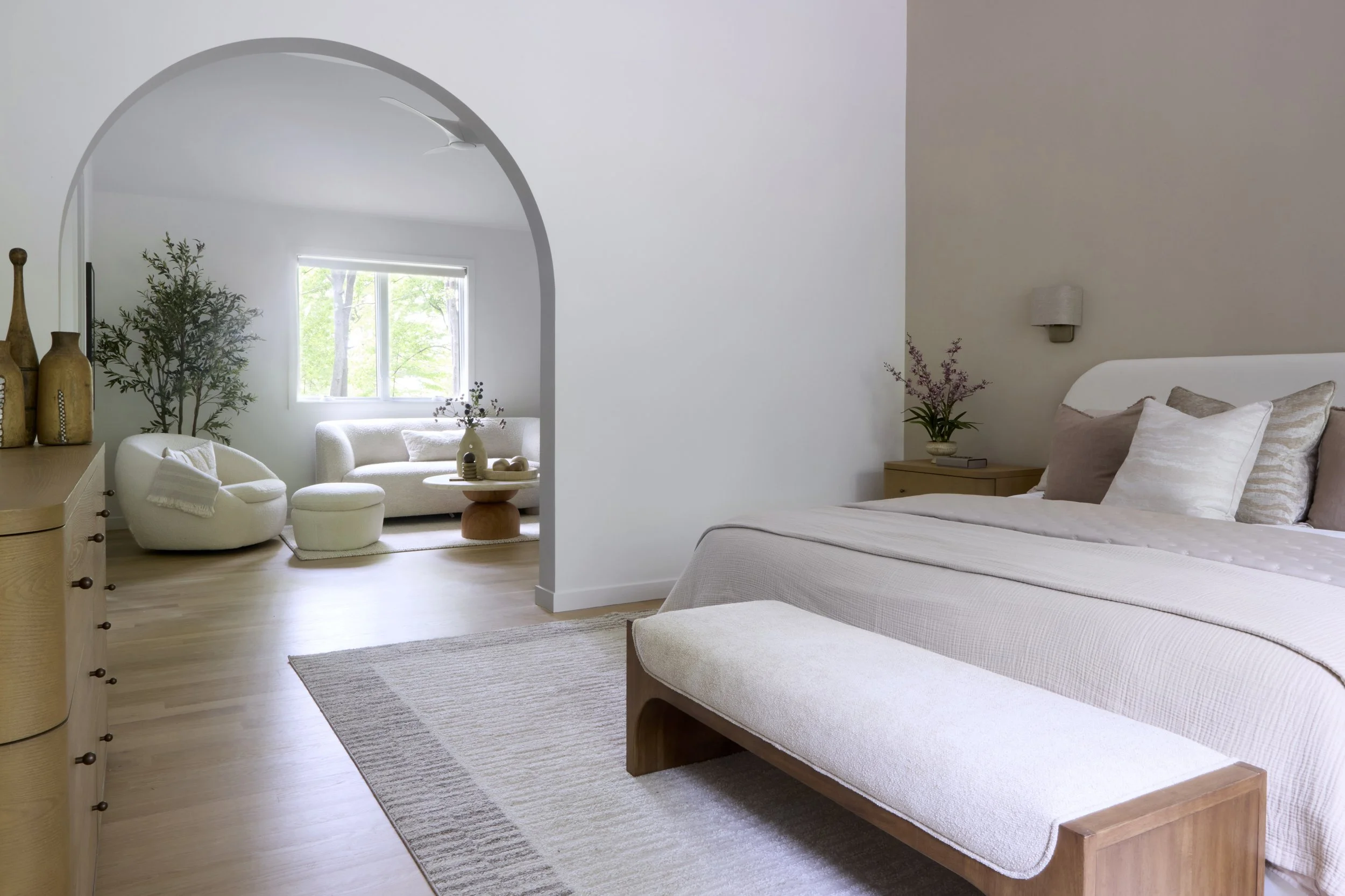 Modern bedroom with a white bed, gray and white pillows, and a floral painting on the wall. Side table with a dark sculpture and books.