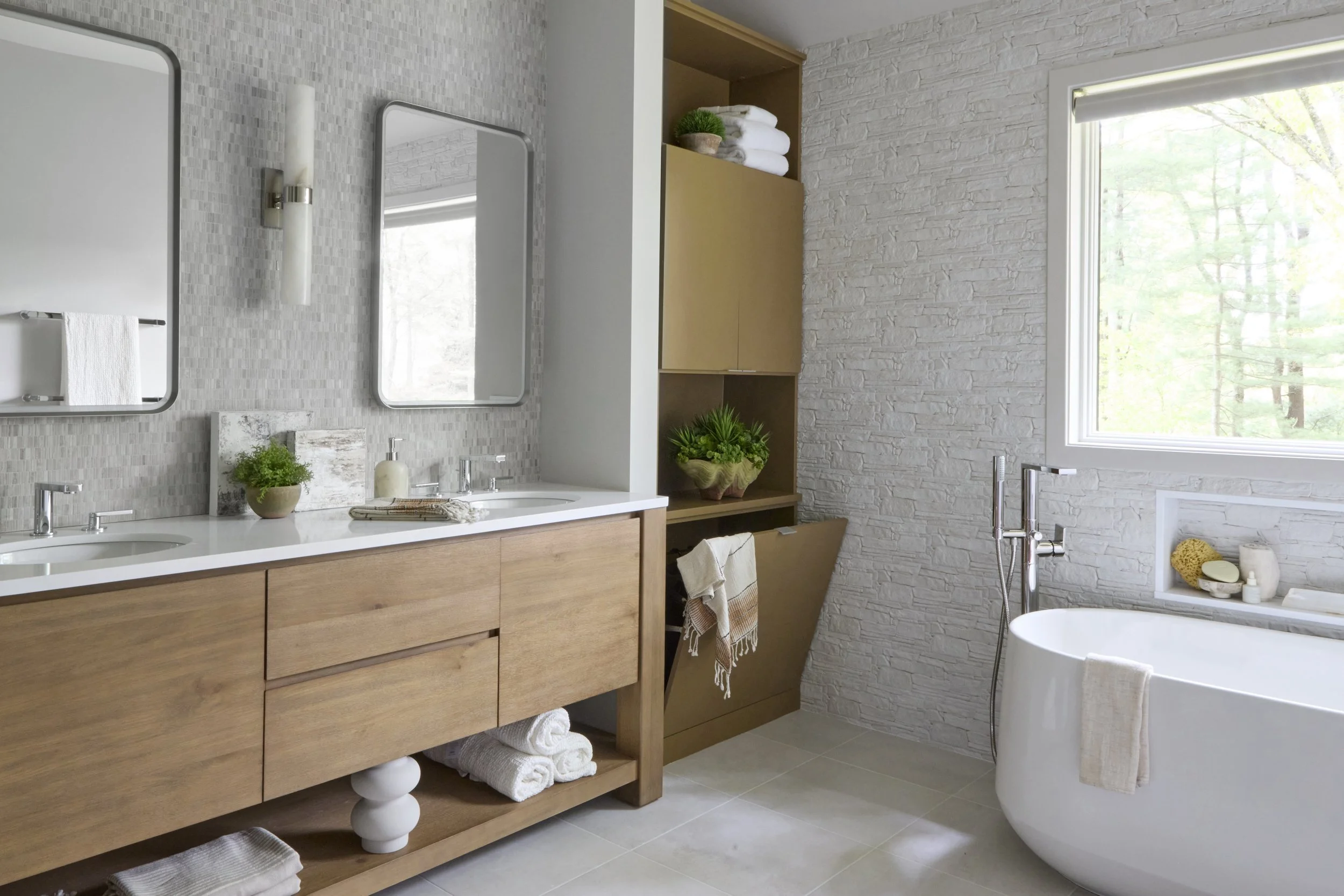 Modern bathroom with freestanding bathtub, wooden vanity, woven rug, wicker basket, hanging lights, and a window.
