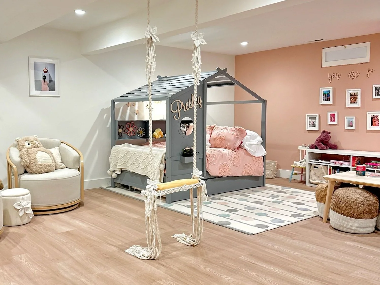 Cozy children's playroom with a small wooden table, woven poufs, a white bookcase with books and bins, wall shelves with framed photos, and a heart-shaped lamp. The room features a polka dot door and a polka dot rug.