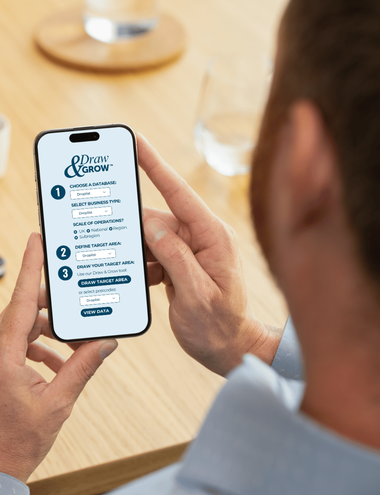 A person is holding a smartphone displaying the Draw & Grow app with options for database selection, business type, scale of operations, target area, and data viewing, while sitting at a wooden table with a glass of water and a round wooden object in the background.