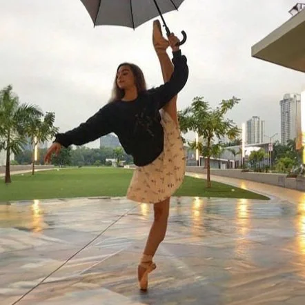 Gabi balances en pointe while holding an umbrella outdoors on a cloudy day.