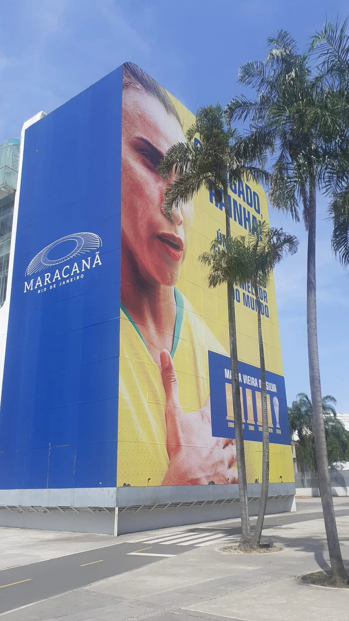 Large billboard advertisement in Rio de Janeiro featuring a woman with an expressive face, holding up one finger in a gesture, with palm trees in the foreground. The billboard is predominantly yellow and blue and includes the logo for 'Maracanã Rio de Janeiro.'