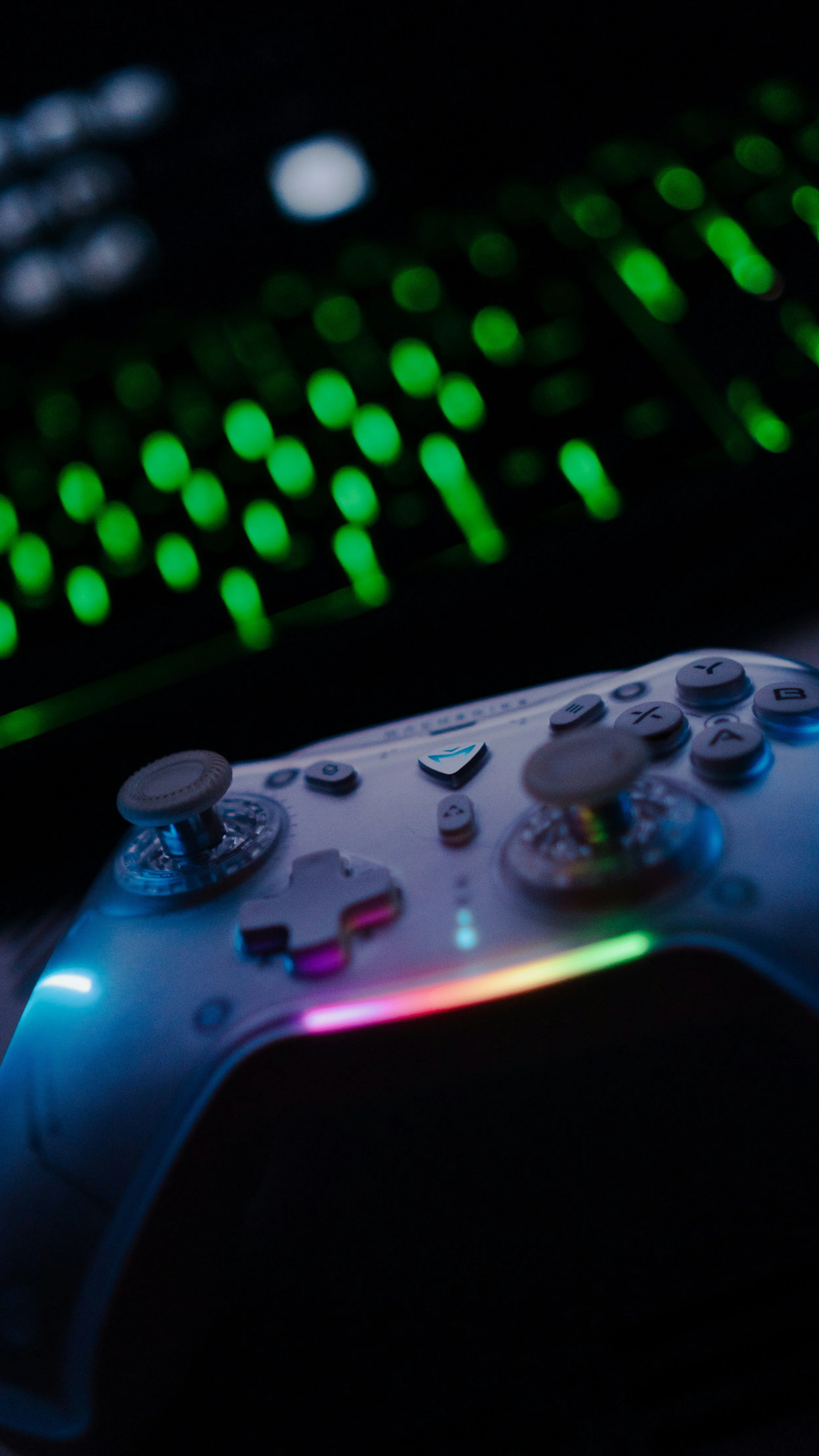 A gaming controller with illuminated colorful lights in front of a blurred gaming keyboard with green backlit keys.