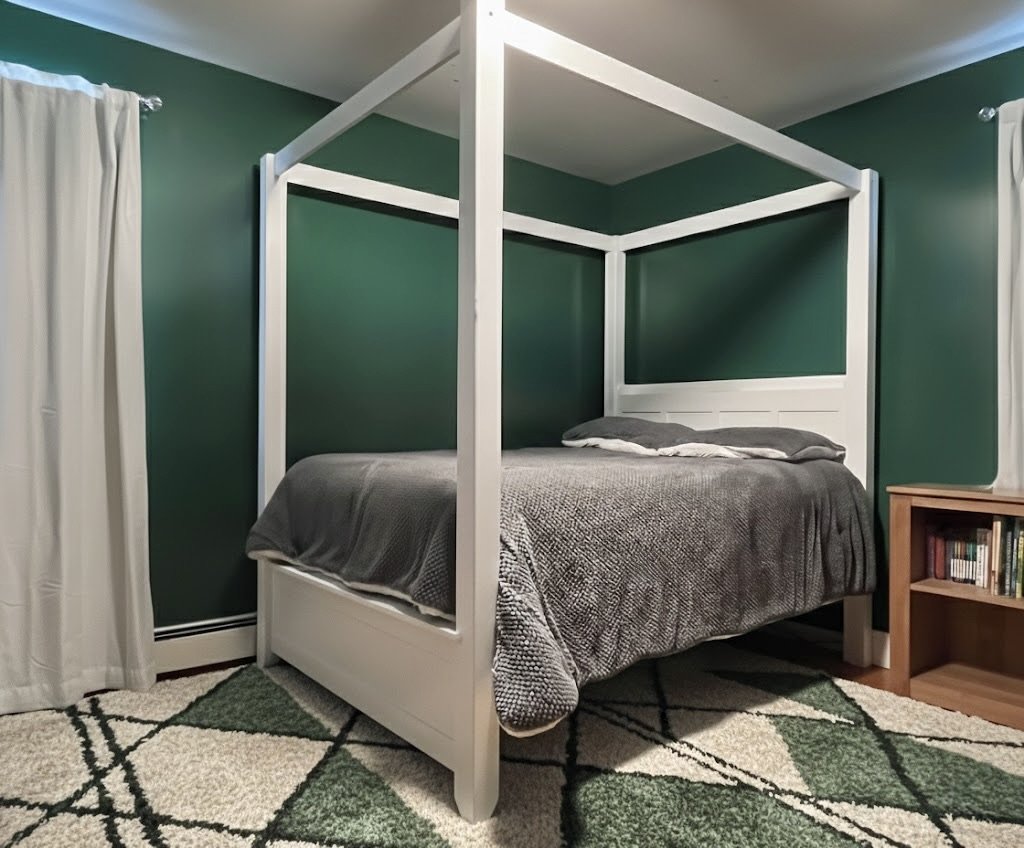 A bedroom with a white four-poster bed, dark gray bedding, green walls, white curtains on the windows, a small wooden bookshelf, and a green and white patterned rug.