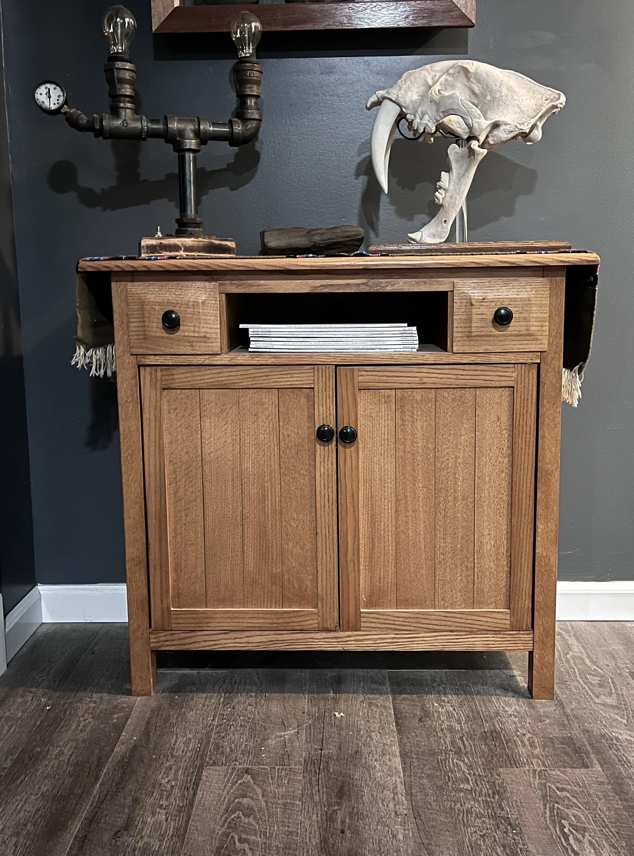 A wooden cabinet with two doors and one open shelf in the middle, topped with a decorative display of a skeleton skull, a piece of wood, a vintage-style wall clock, and a chandelier made of pipe fittings with light bulbs. The cabinet is against a dark gray wall and has a wooden floor.