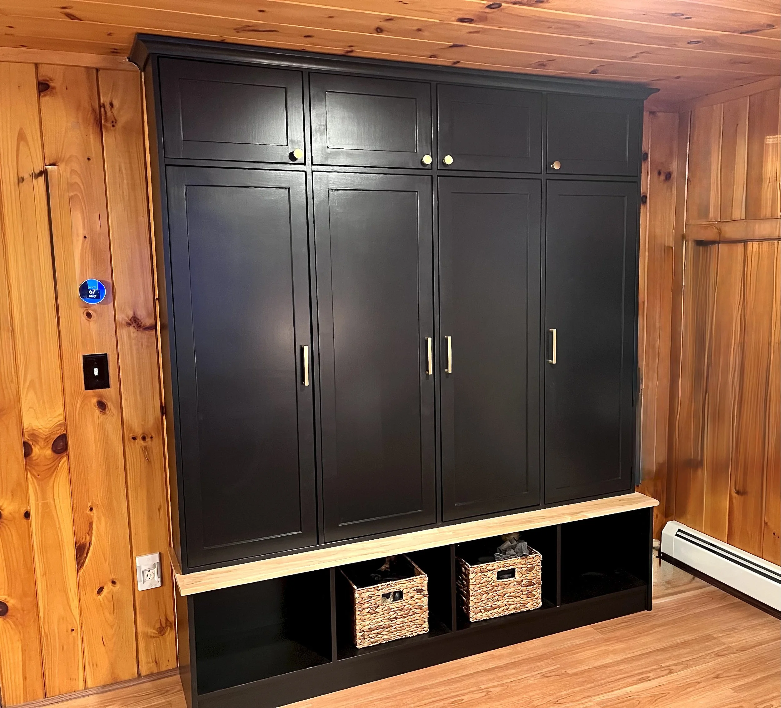 A large black wardrobe with three doors and gold handles, sitting on a wooden shelf with three woven baskets beneath. The room has wood-paneled walls and flooring.