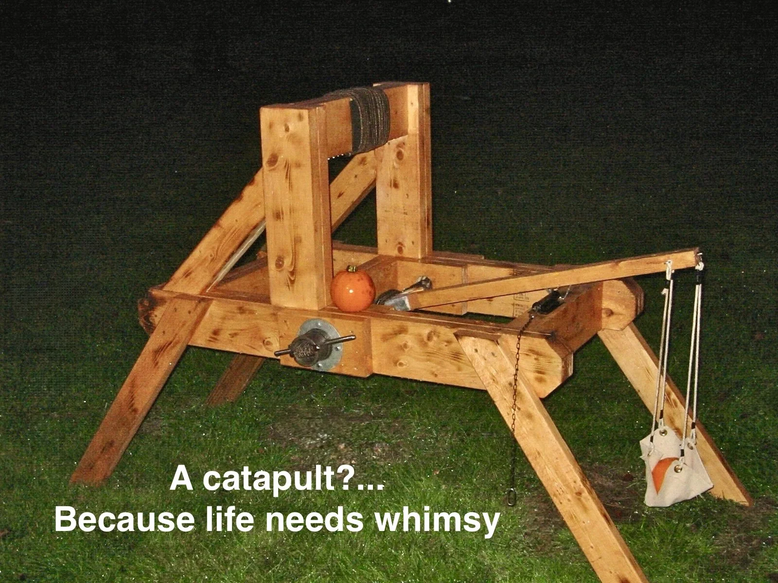 A wooden catapult with a pumpkin placed on it, set against a dark outdoor background at night. The catapult has adjustable parts, a sling with a small bag, and a metal lever for launching.