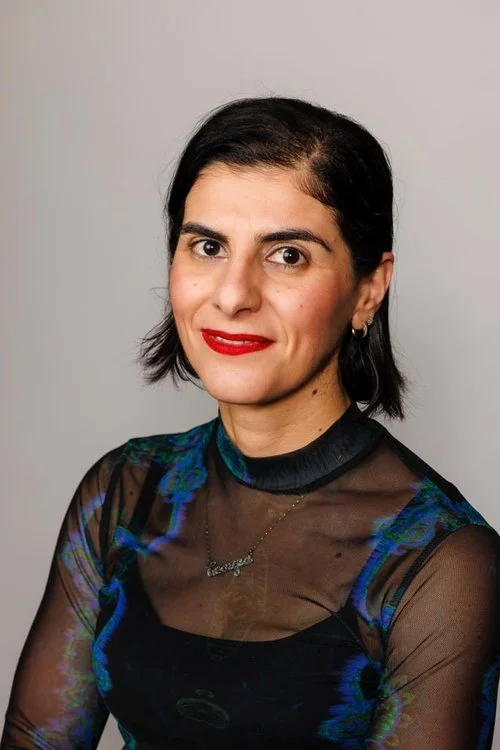 A woman with short dark hair and red lipstick wearing a dark, sheer top with blue accents and a necklace against a plain light background.