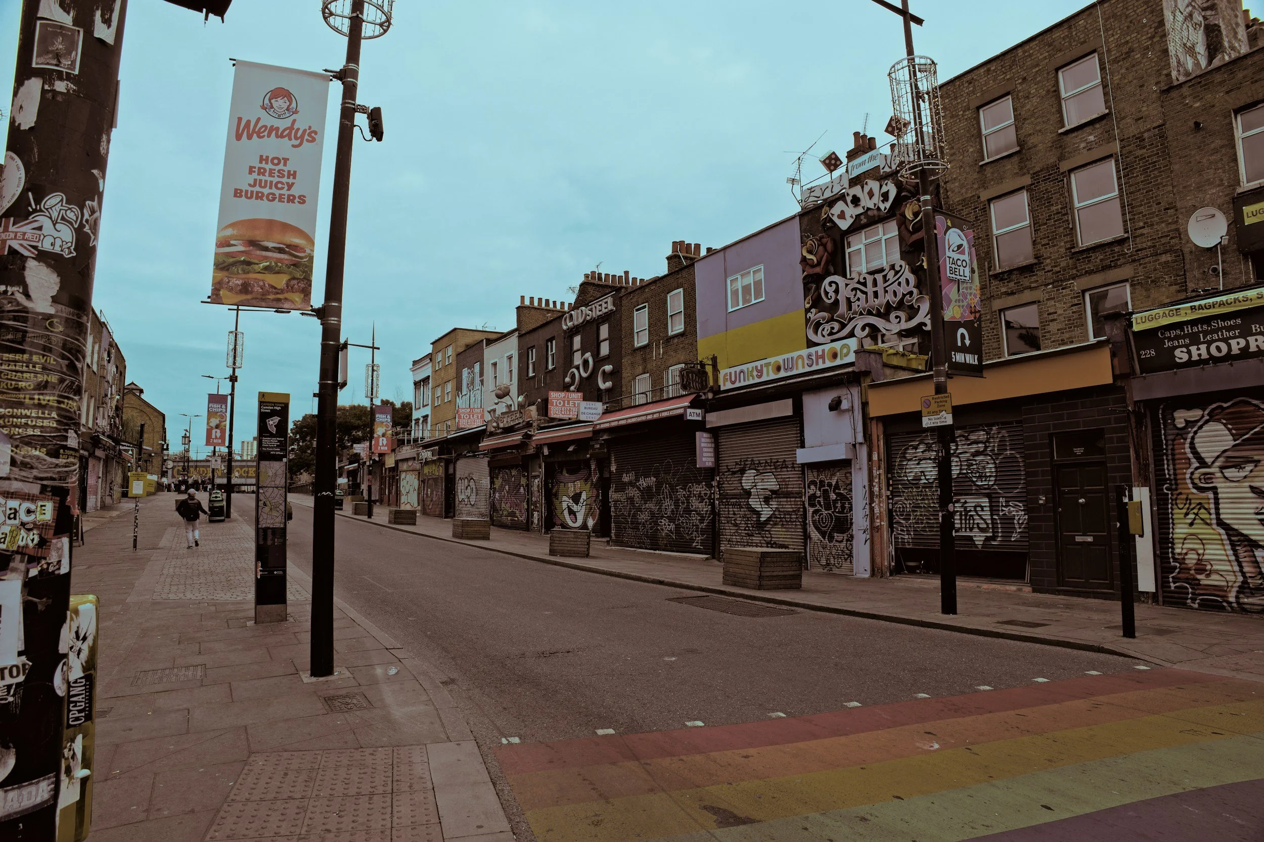 Empty city street with closed storefronts, graffiti-covered shutters, street signs, and a Wendy's banner overhead, with a person walking down the sidewalk.