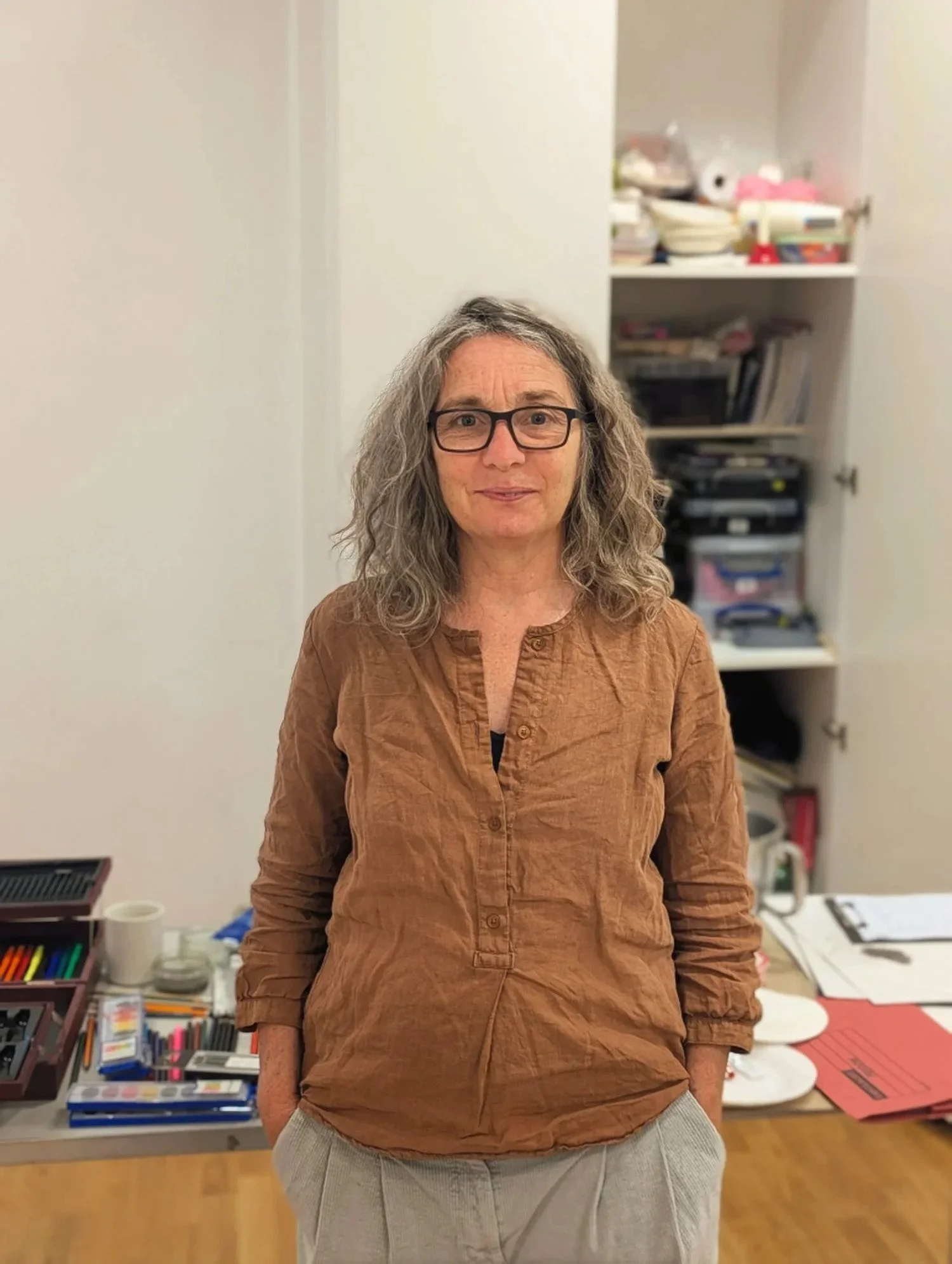 A woman with curly gray hair and glasses stands in a room with cluttered shelves and a table with various office supplies.