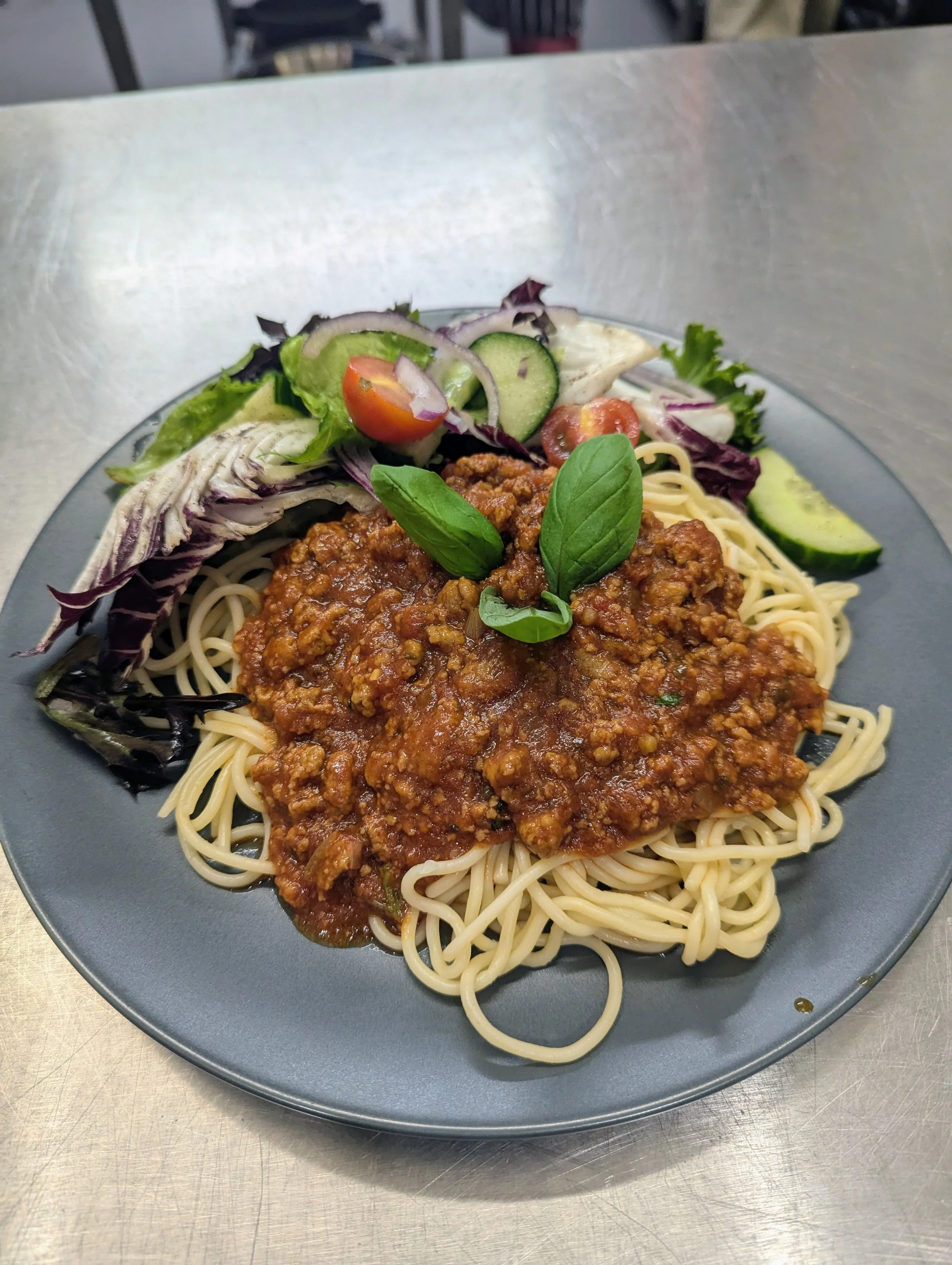 Plate of spaghetti with meat sauce, served with mixed salad including cherry tomatoes, cucumber slices, and lettuce, garnished with fresh basil leaves.