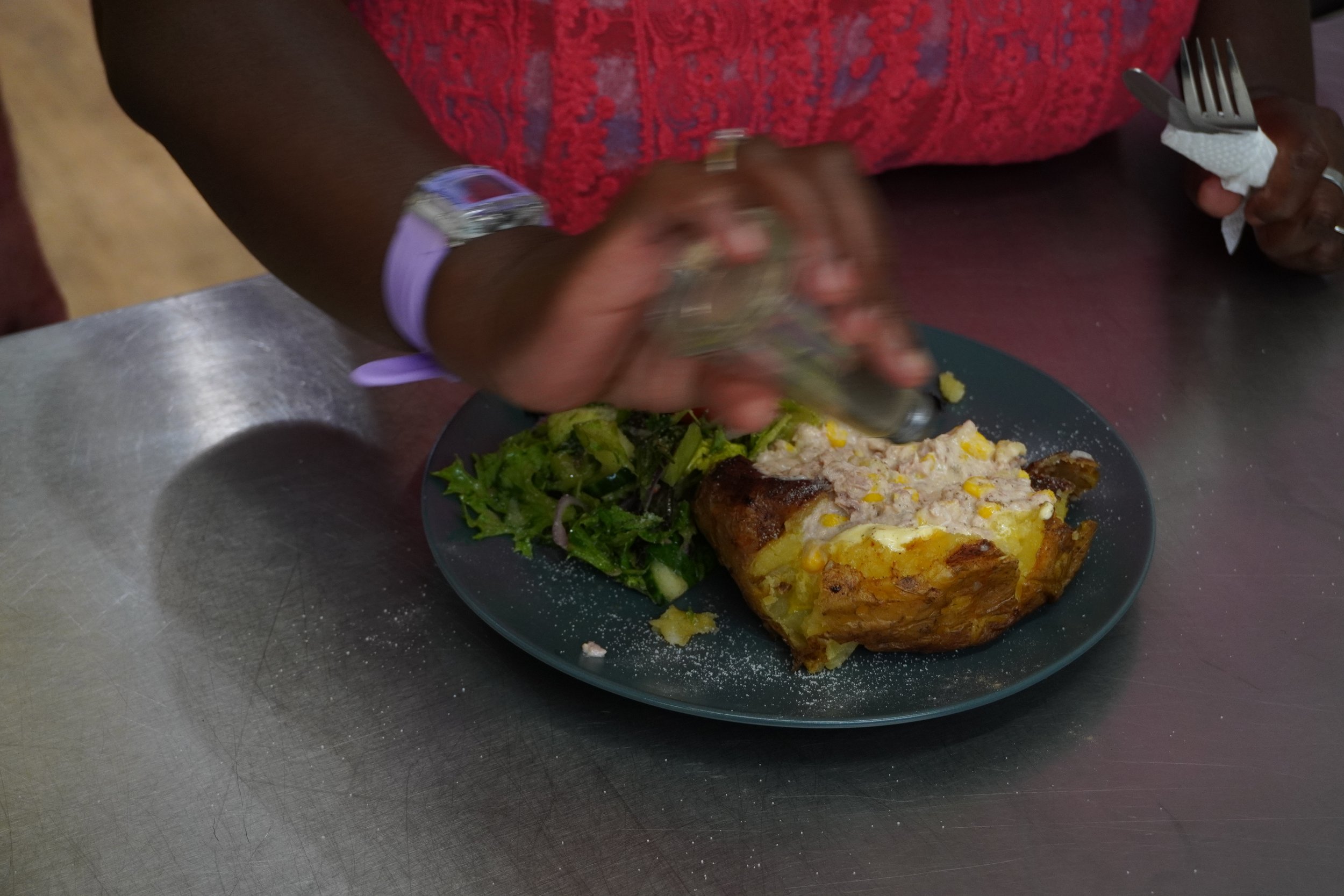 A woman shakes pepper onto a jacket potato with tuna sweetcorn and a side of lettuce.
