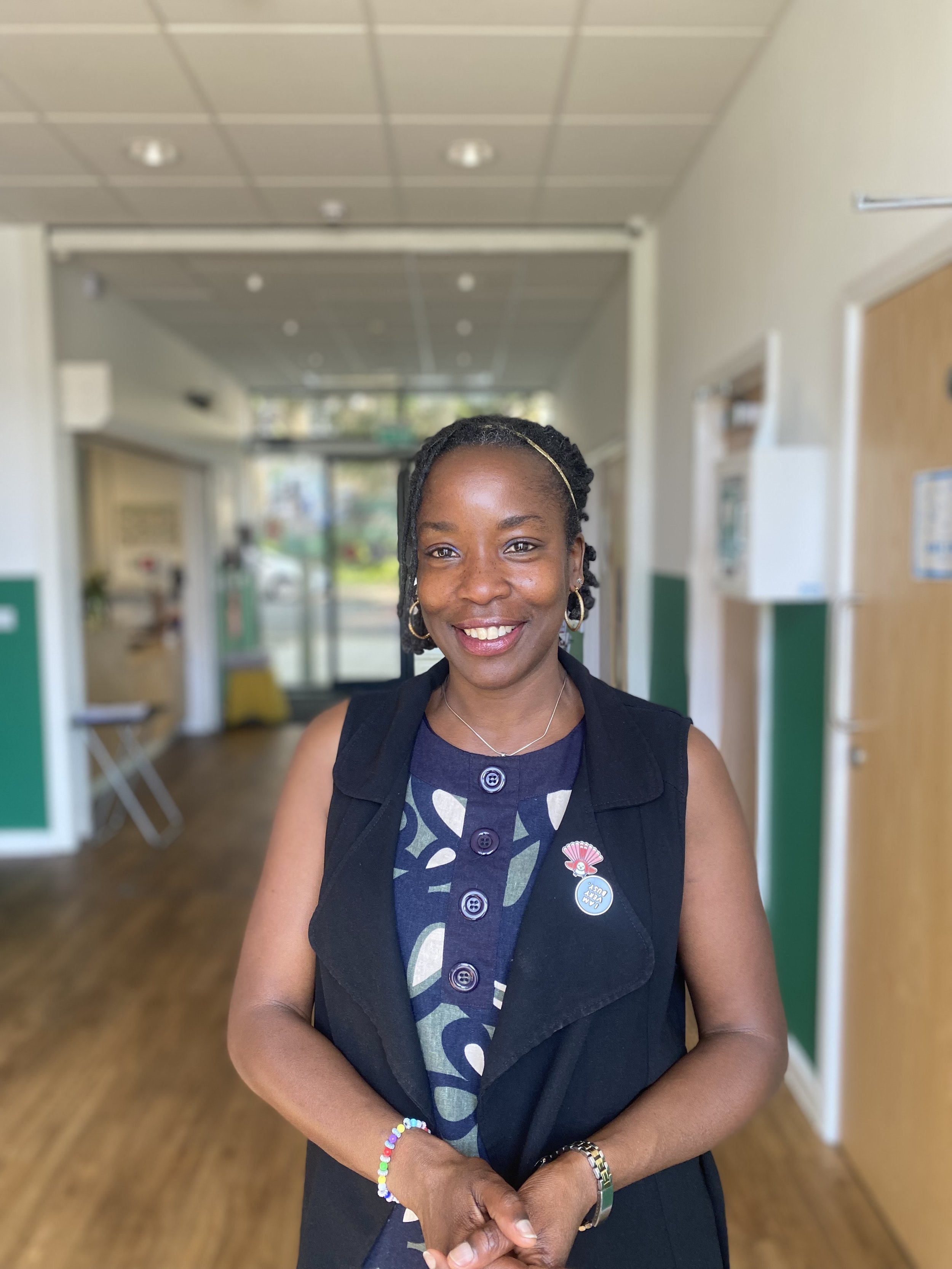 A smiling Black woman with braided hair wearing a sleeveless black vest over a patterned dress, standing inside a building with a wooden floor and a ceiling with recessed lighting.