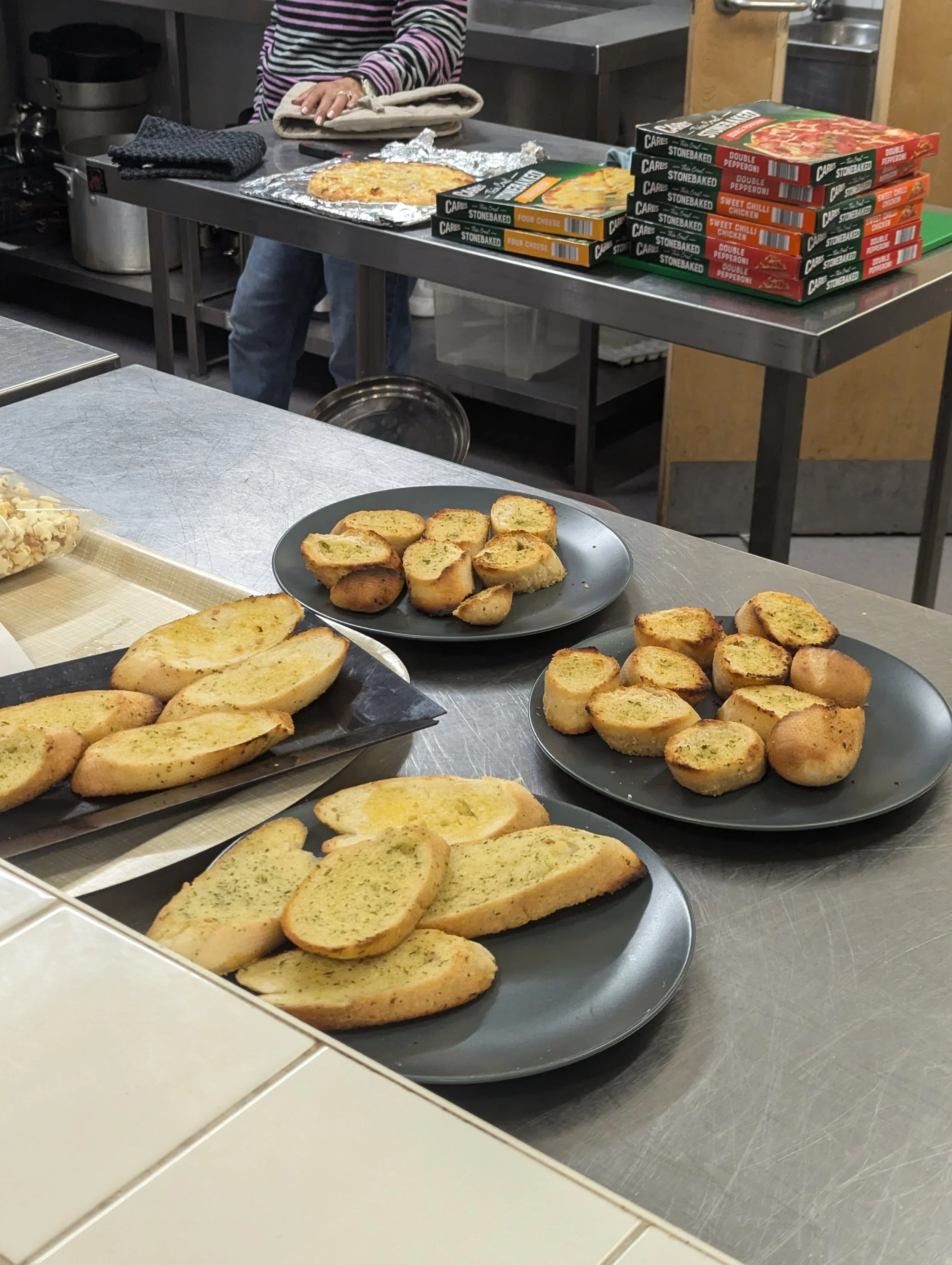 Plates of garlic bread on a stainless steel kitchen counter with frozen pizza boxes stacked in the background.