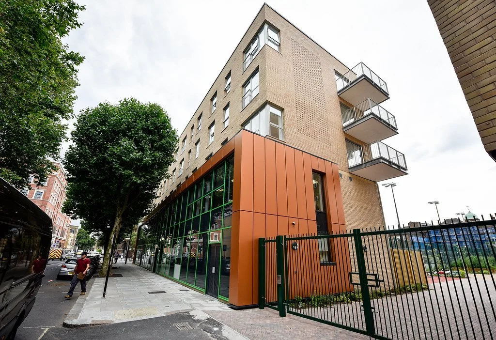 A modern multi-story building with a brick facade and large glass windows, featuring balconies on one side, located on a city street with trees, pedestrians, and a sports field in the background.