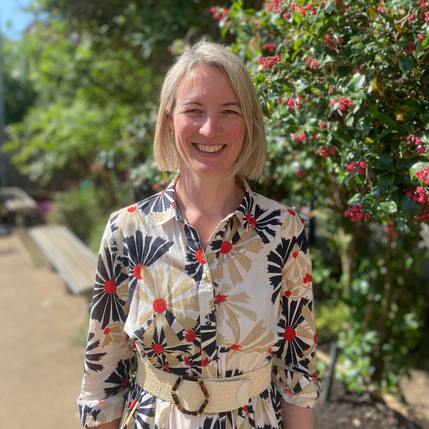 A woman with chin-length blonde hair, smiling, standing outdoors in front of greenery with pink flowers. She is wearing a patterned dress with a beige belt.