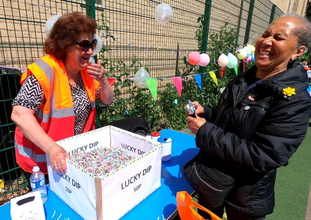 Two women laughing at a carnival booth with a box labeled "Lucky Dip" filled with candies. One woman wearing sunglasses and an orange safety vest, the other holding a wrapped candy. Colorful balloons and flags decorate the background.