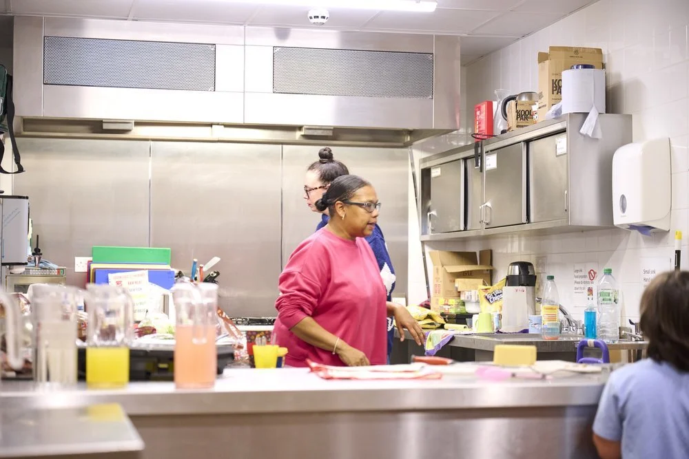 Kitchen with three women preparing food or drinks. Counter with cups, bottles, and kitchen equipment.