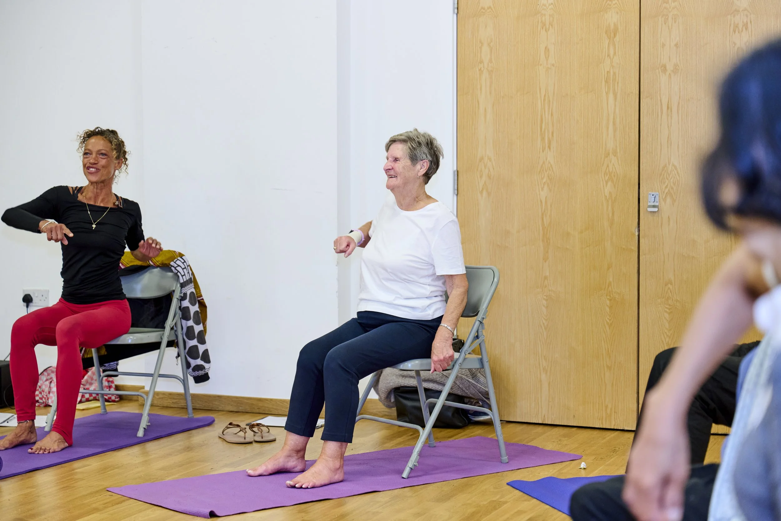 Two women sitting on chairs and stretching on yoga mats in a room, with a wooden door, and a person partially visible on the right side.