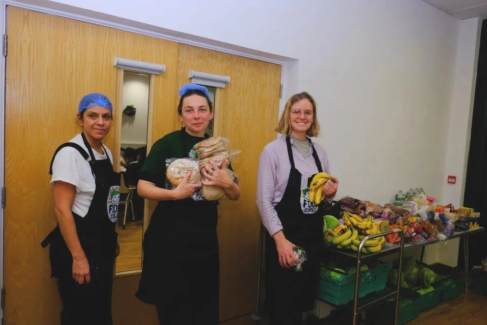 Three women wearing aprons and hair caps standing in front of a table of bananas and other food items, smiling.