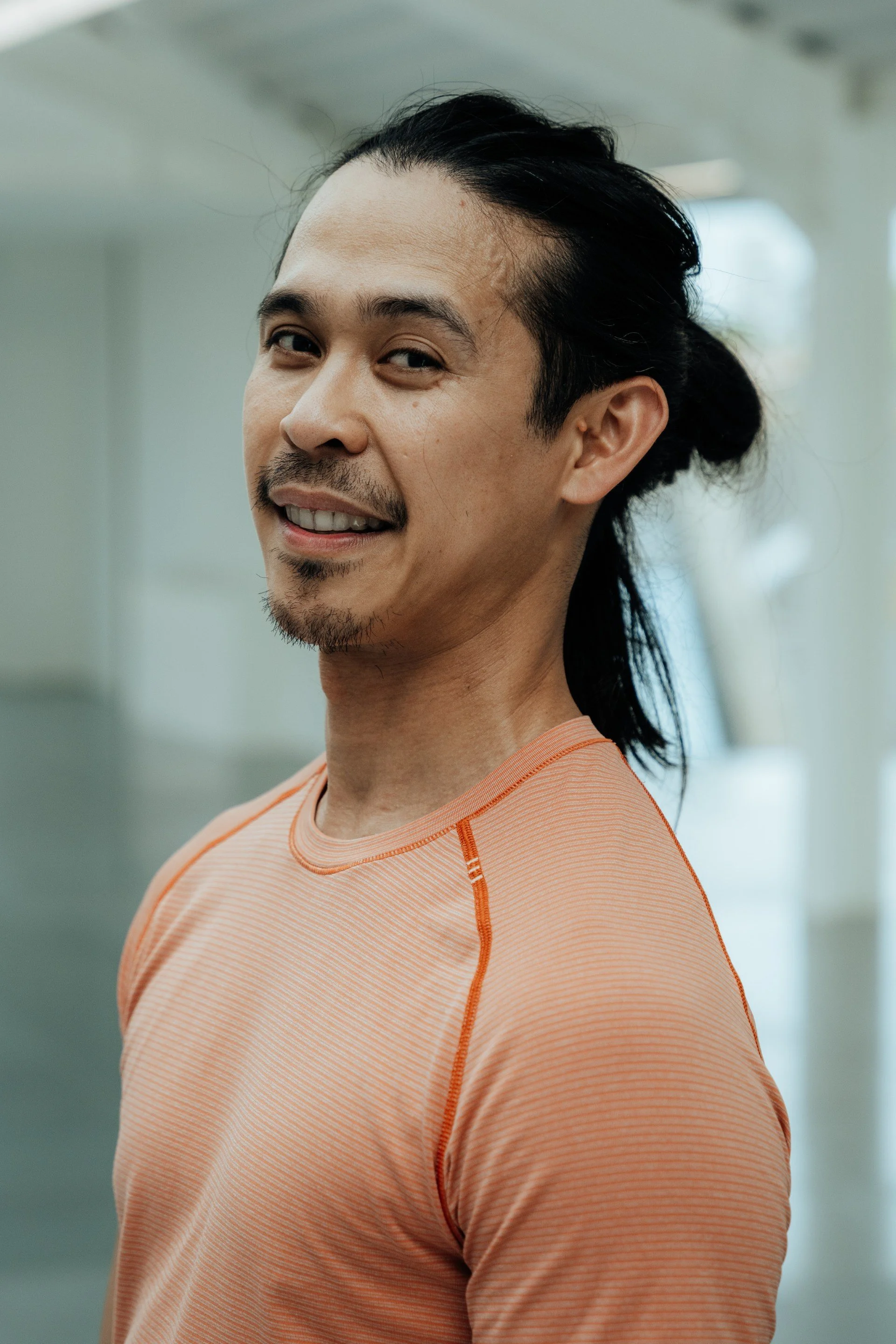 A man with long black hair tied back, light facial hair, smiling slightly, wearing an orange athletic shirt.