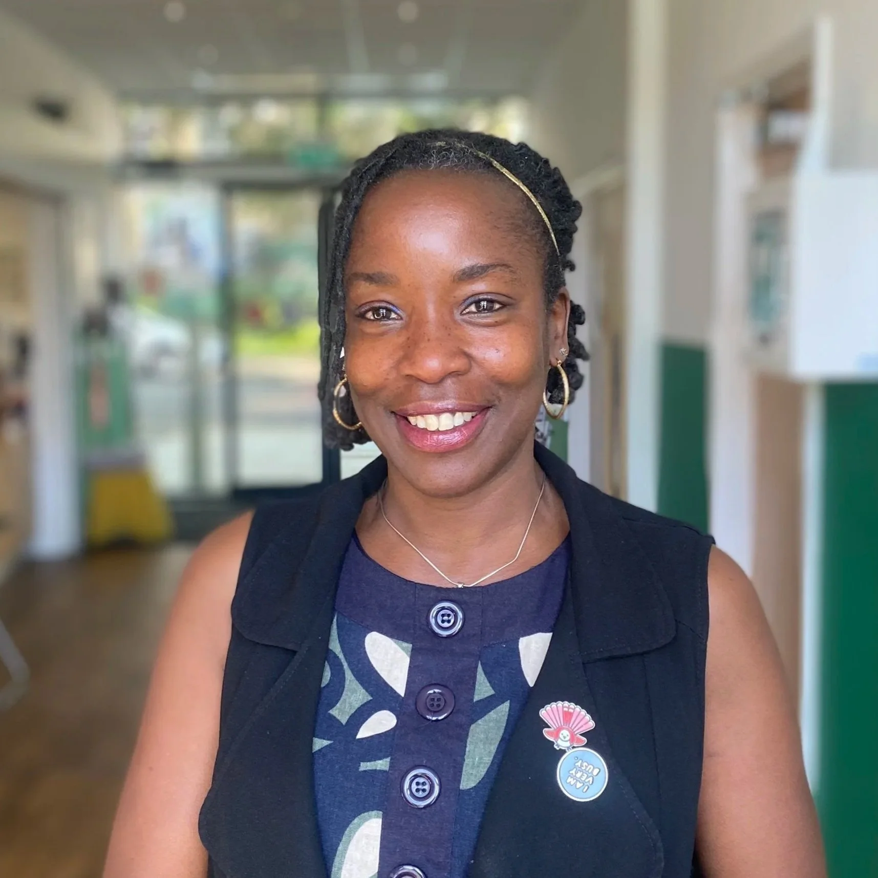 A woman smiling inside a building, wearing a navy blue dress with buttons, a black vest, and jewelry, with pins on her vest.