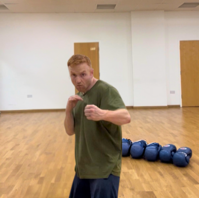 A man in a green shirt posing in a boxing stance in a gym with blue kettlebells lined up on the floor behind him.