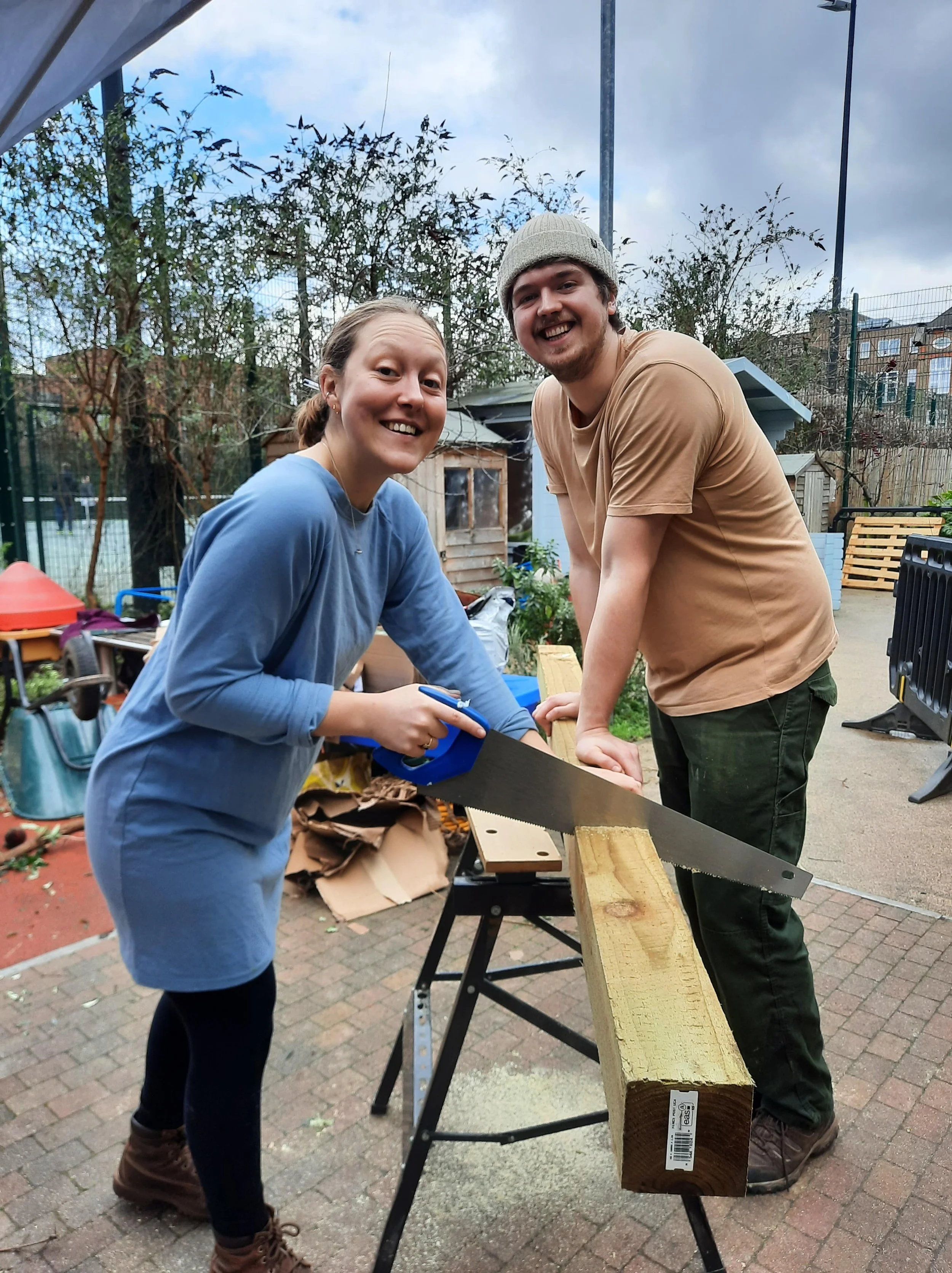 Two people smiling and working together on a woodworking project outdoors, using a saw on a wooden plank.