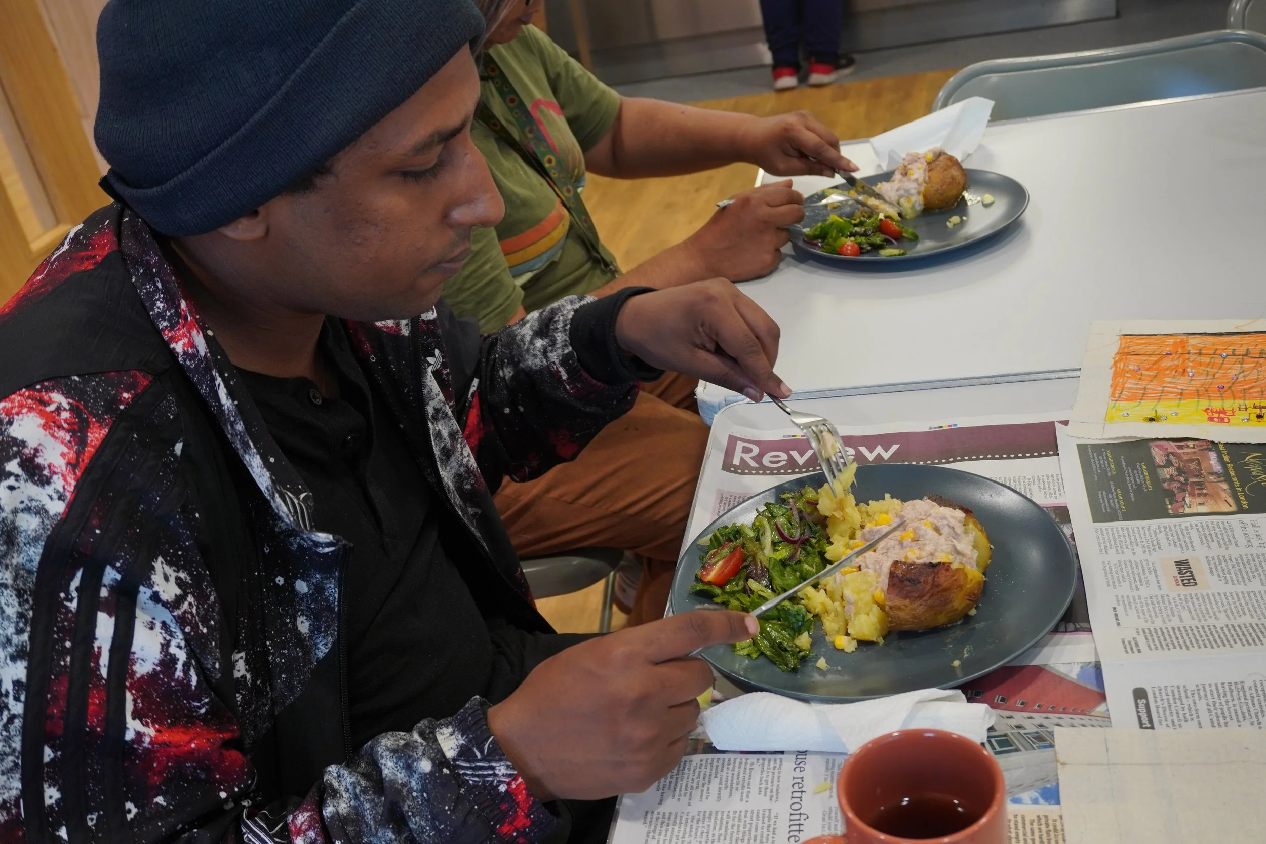 A young man in a beanie and sports jacket eats a jacket potato with tuna sweetcorn with a side of lettuce and cherry tomatos.