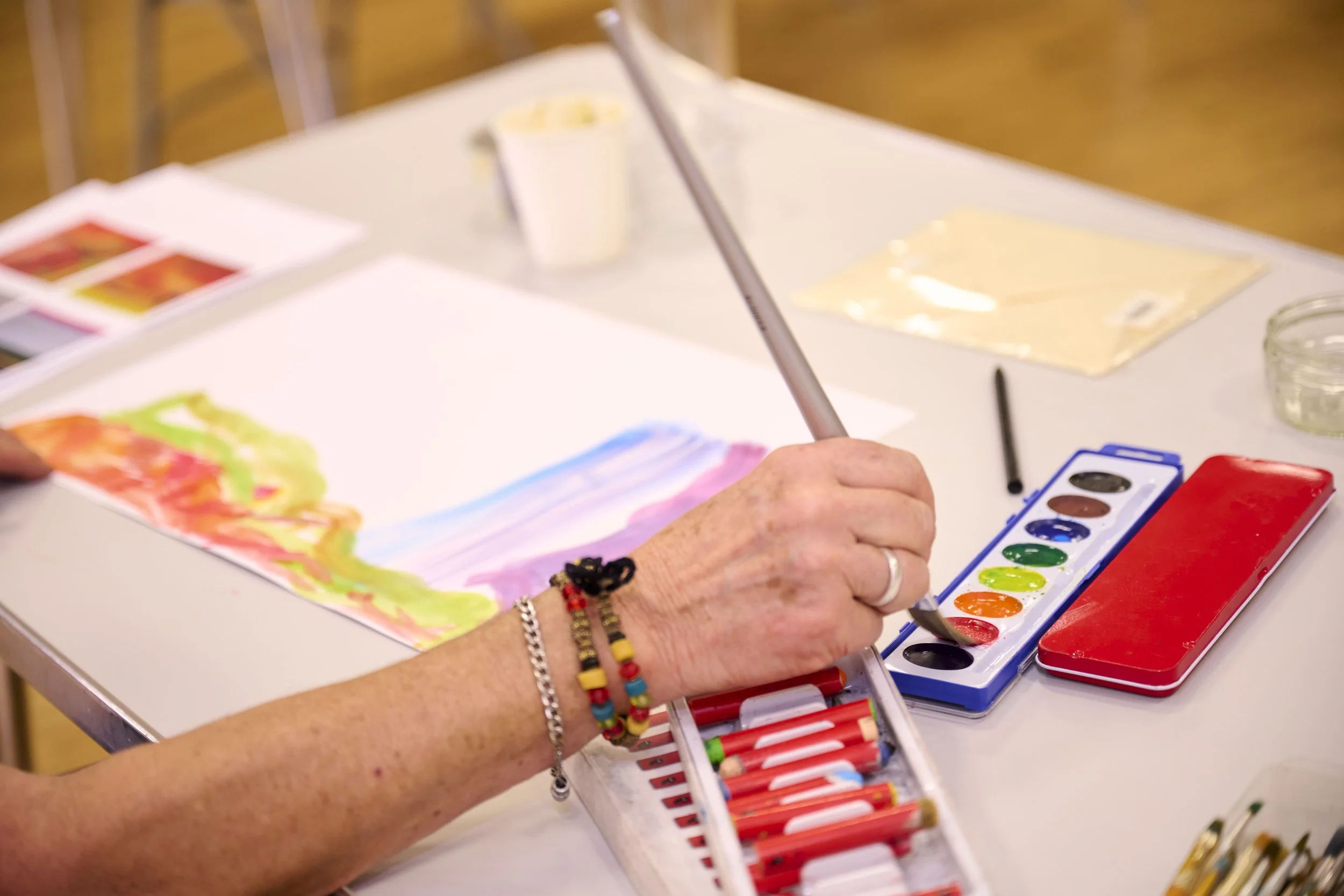 Person creating watercolor painting with red, orange, yellow, green, blue, and purple colors on paper, with watercolor paints and brushes on the table.