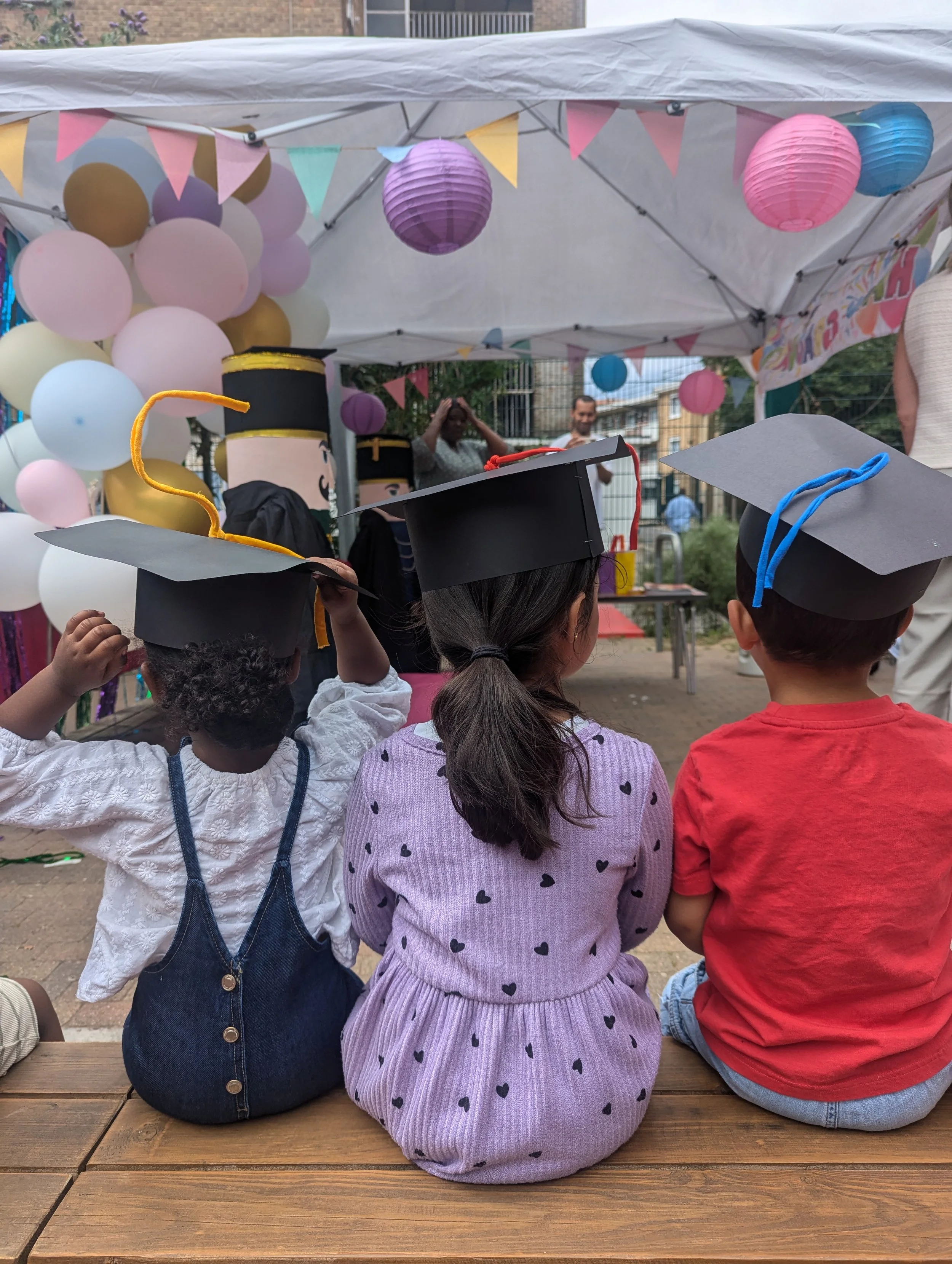 Three children wearing graduation caps and gowns sitting on a bench at an outdoor celebration with balloons and decorations.