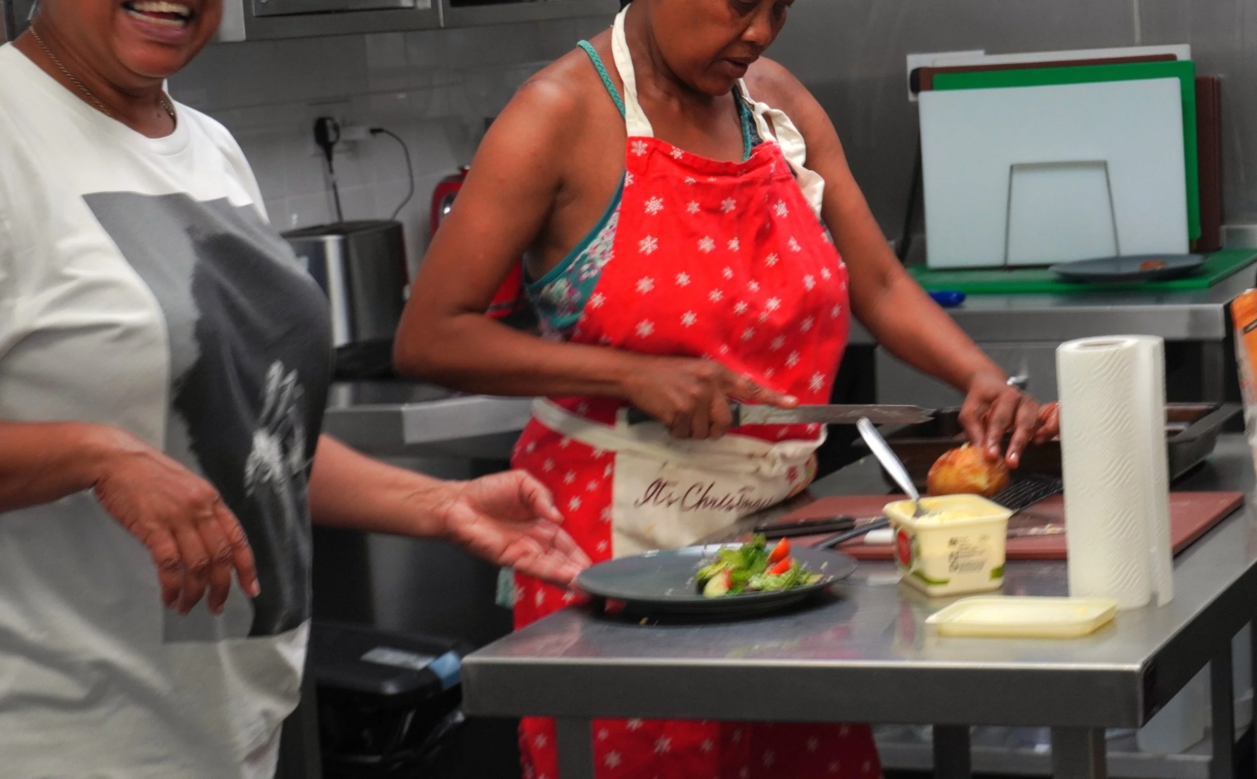 Two women prepare a plate, one cutting a potato and the other putting the plate with salad down and laughing.