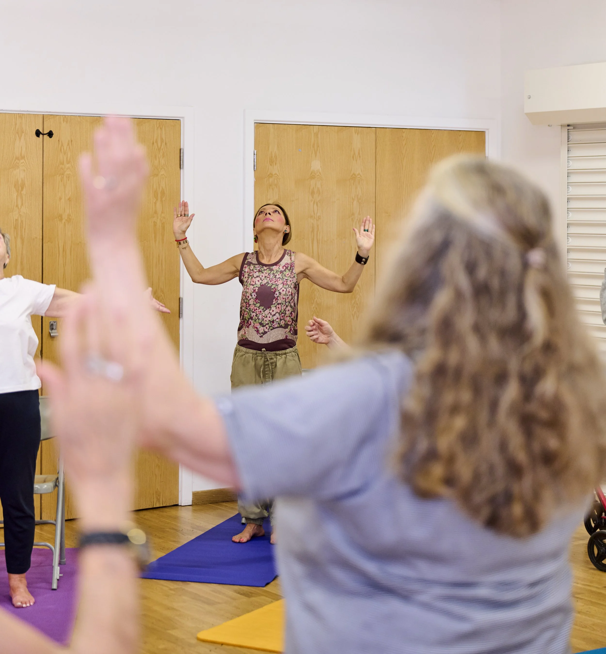 A woman standing on a yoga mat with her arms raised, leading a group exercise or yoga class in a room with wooden doors and white walls.