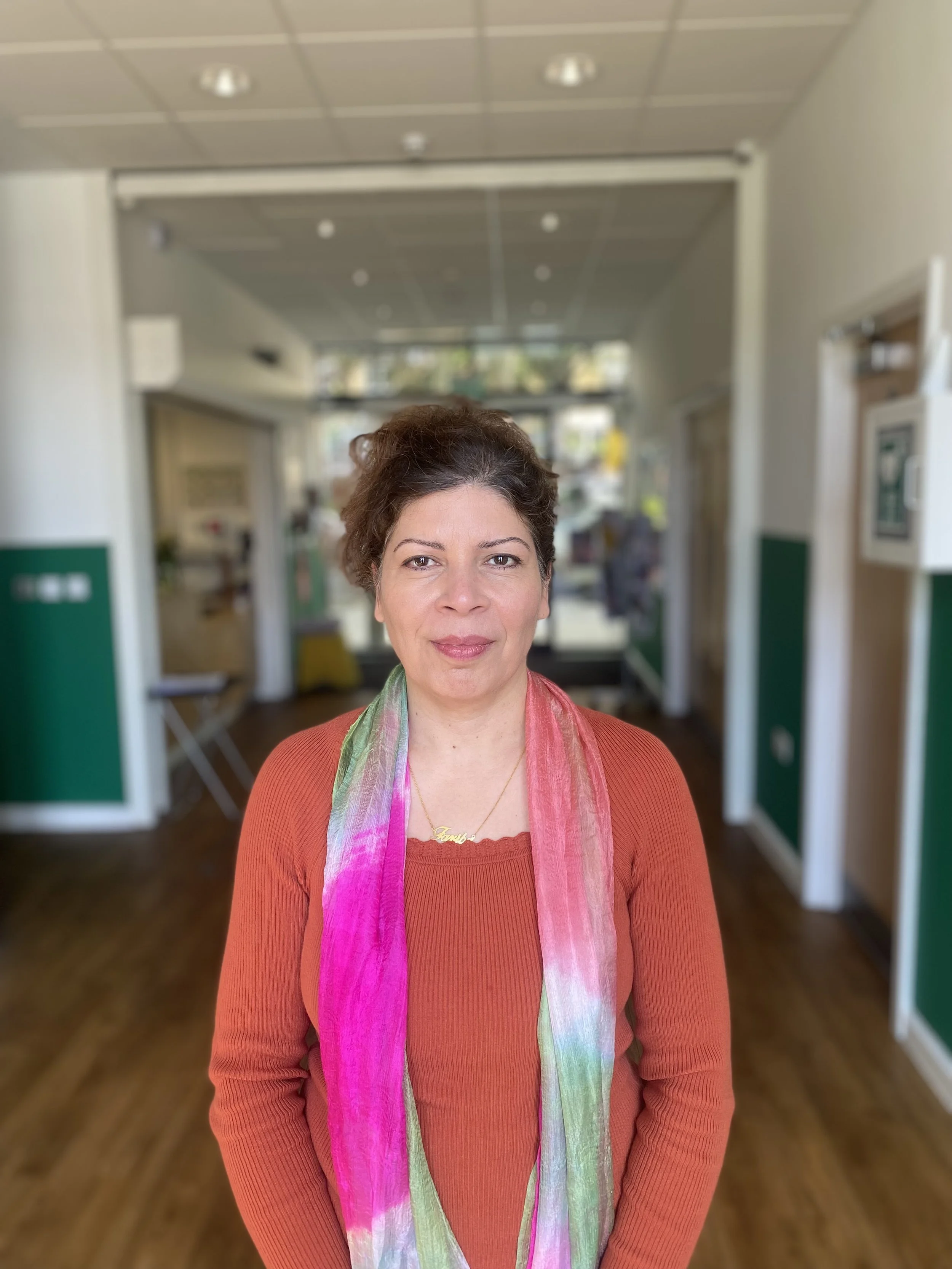 A woman standing indoors, wearing an orange sweater and a colorful scarf, with a blurred background of a lobby or reception area.