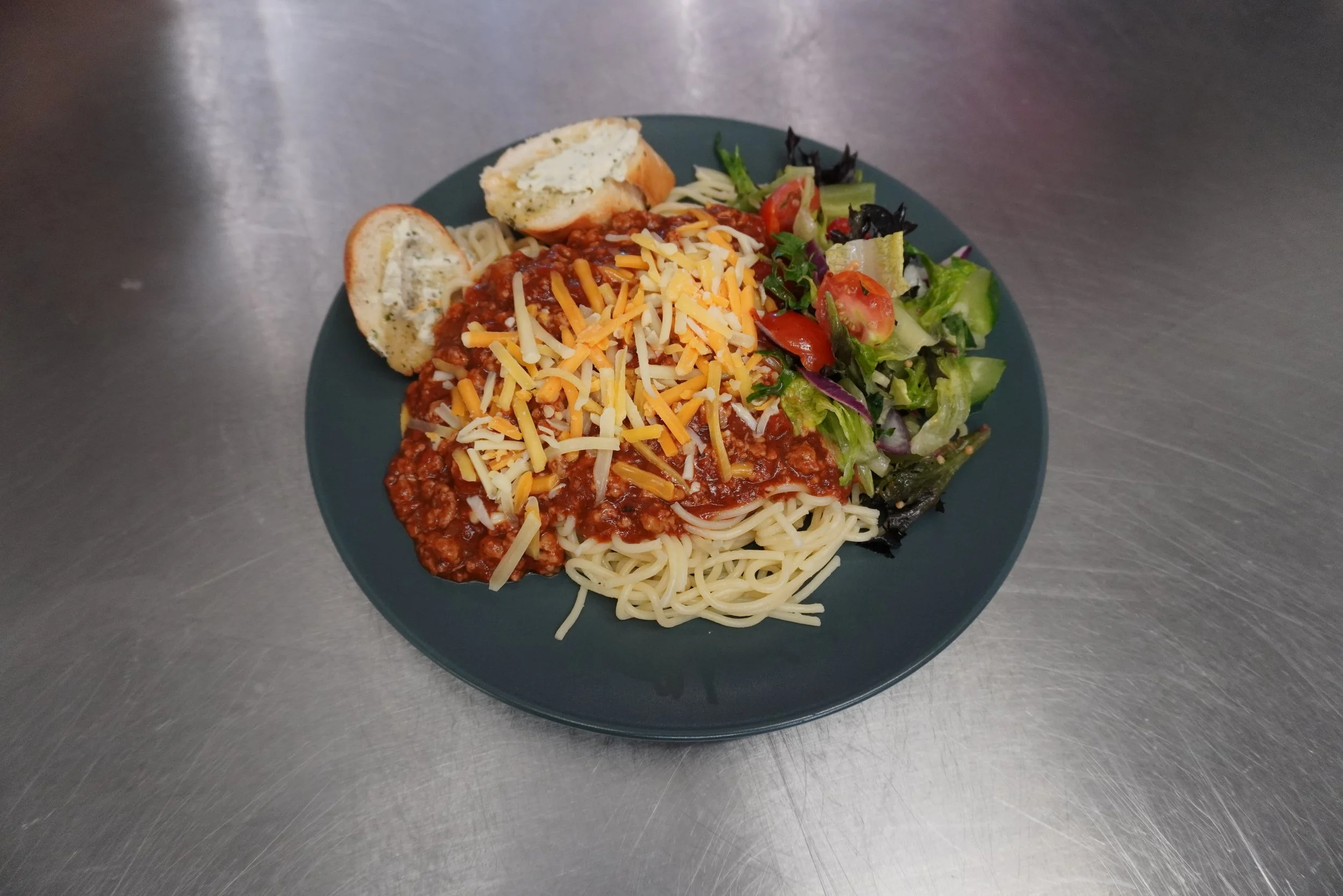 Plate of spaghetti with meat sauce topped with white and yellow cheese shreds, served with mixed salad including cherry tomatoes and lettuce, and two slices of garlic bread with butter.