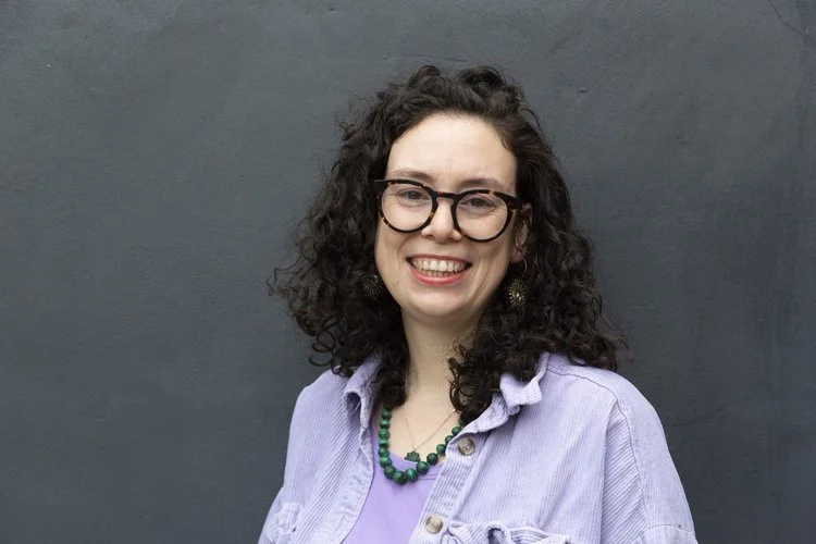 A woman with curly dark hair, glasses, and earrings, smiling in front of a gray wall, wearing a lavender shirt and a beaded necklace.