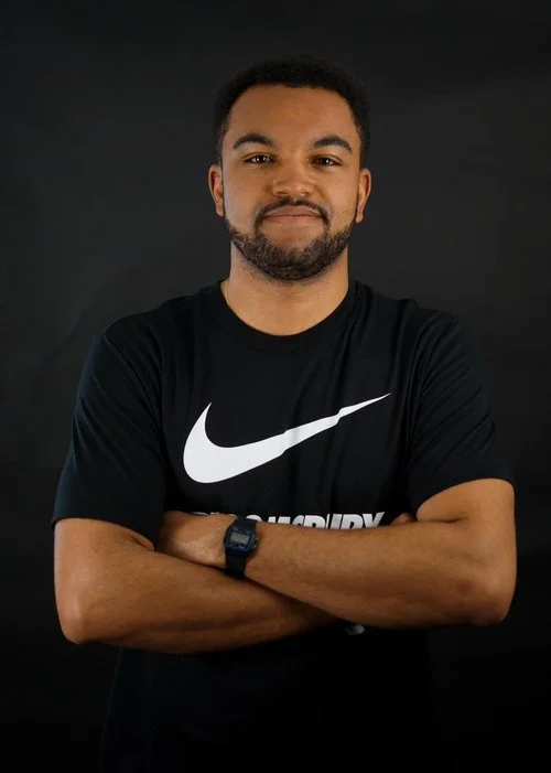 A young man with a beard and short curly hair, wearing a black Nike T-shirt with a white swoosh logo, standing with arms crossed against a black background.