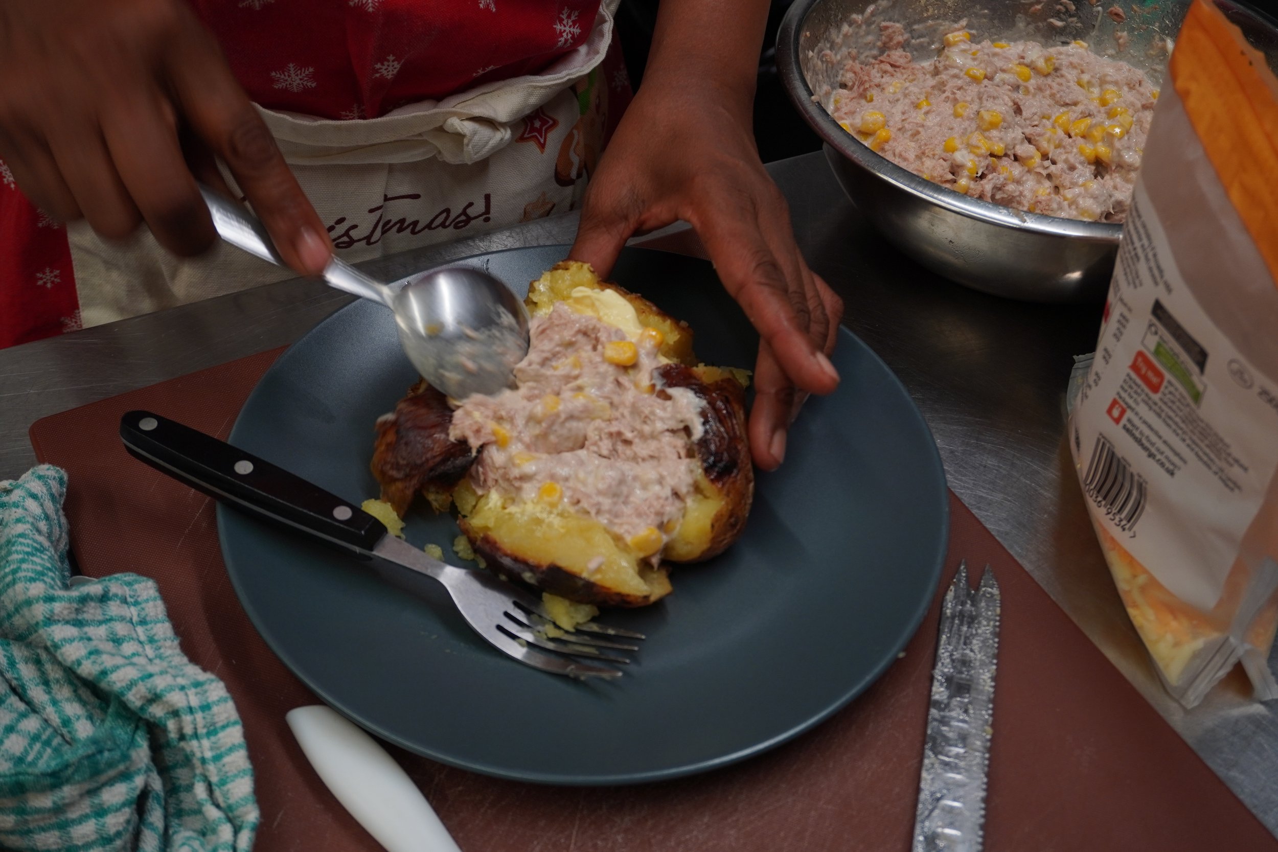Hands prepare a jacket potato with tuna sweetcorn on a navy plate.