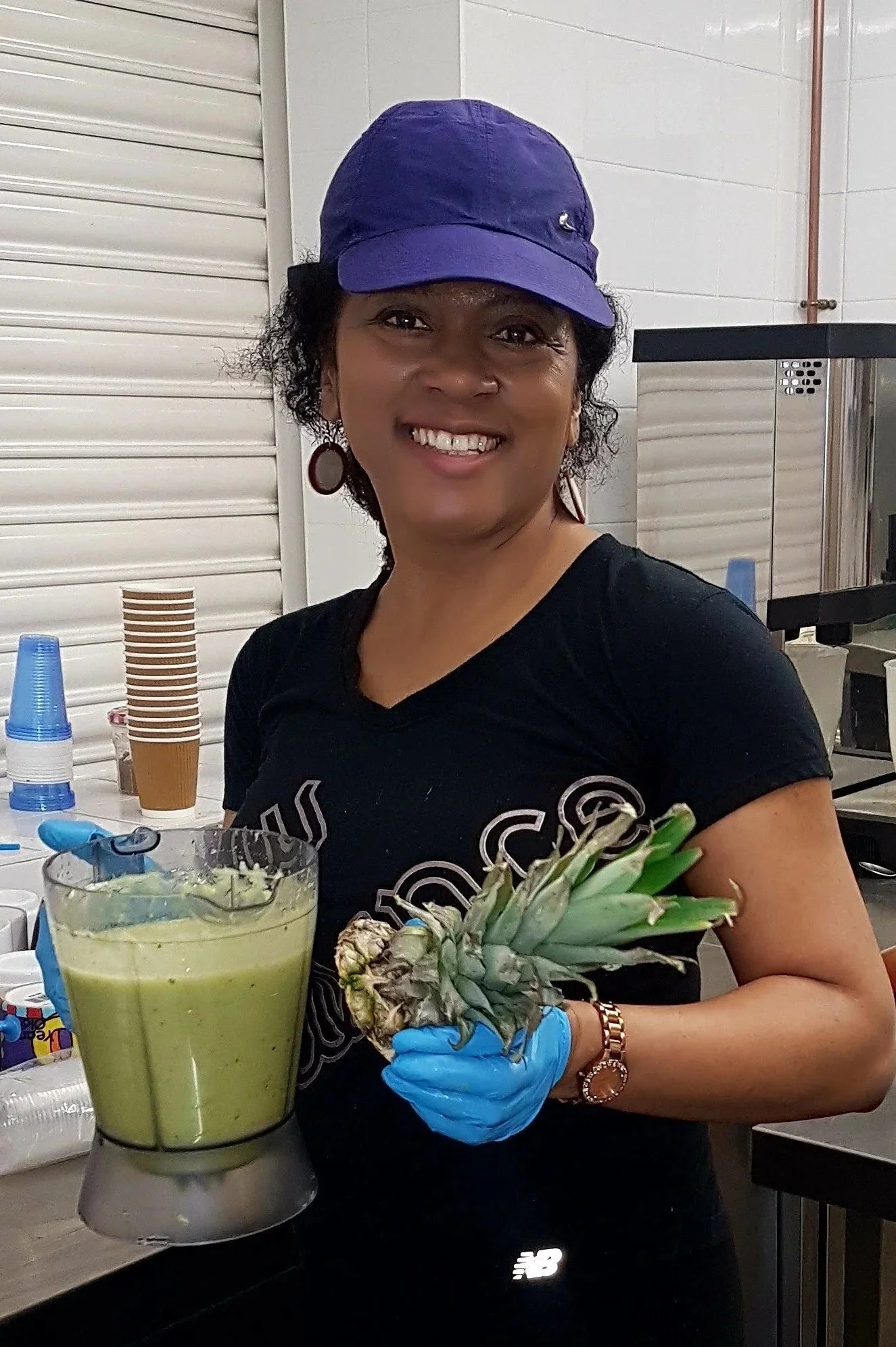 A woman smiling while holding a pineapple and wearing a black shirt, purple cap, and blue gloves, standing in a kitchen or food preparation area.
